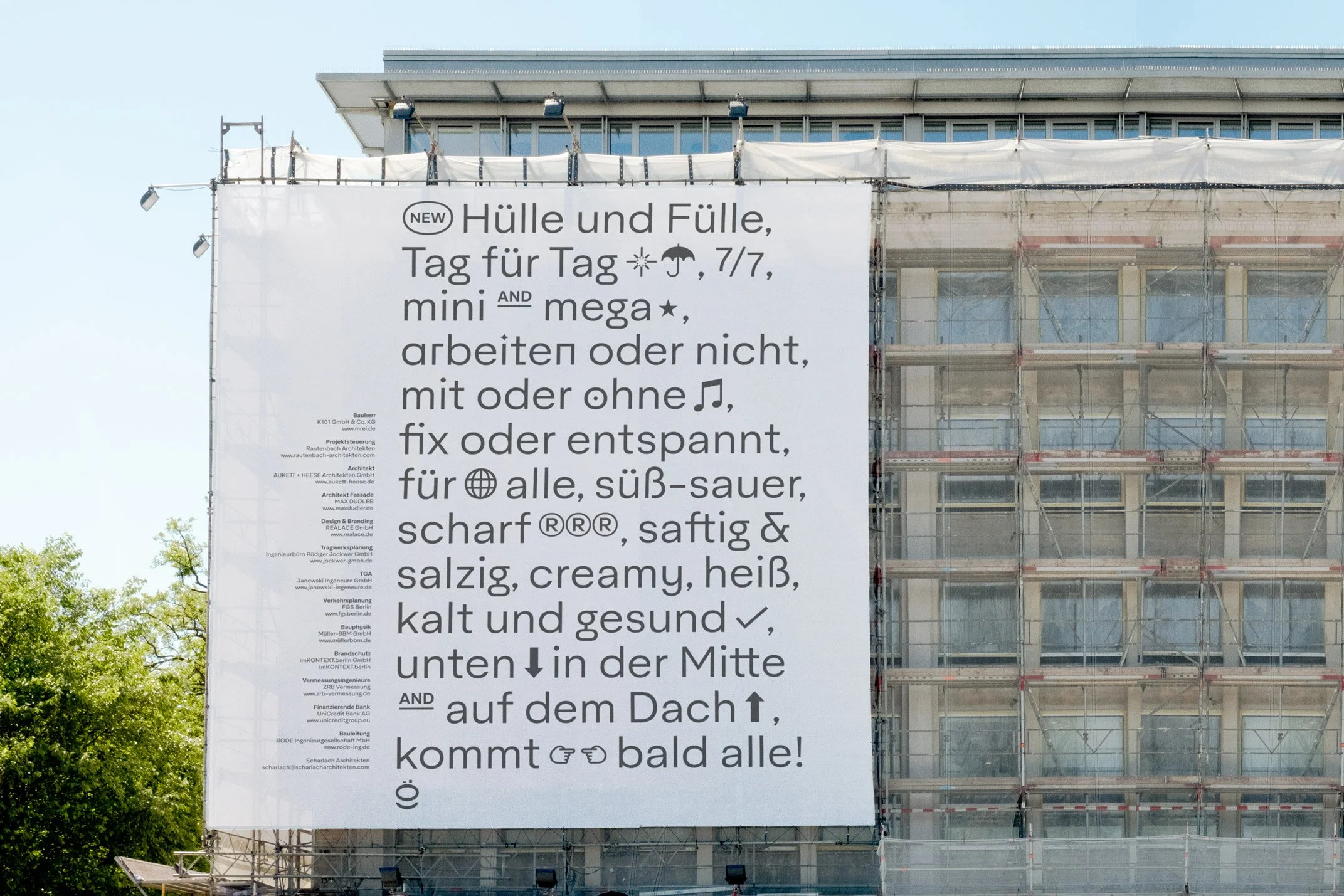 Large white banner with German text and symbols hung on a building under construction, with scaffolding and trees visible.