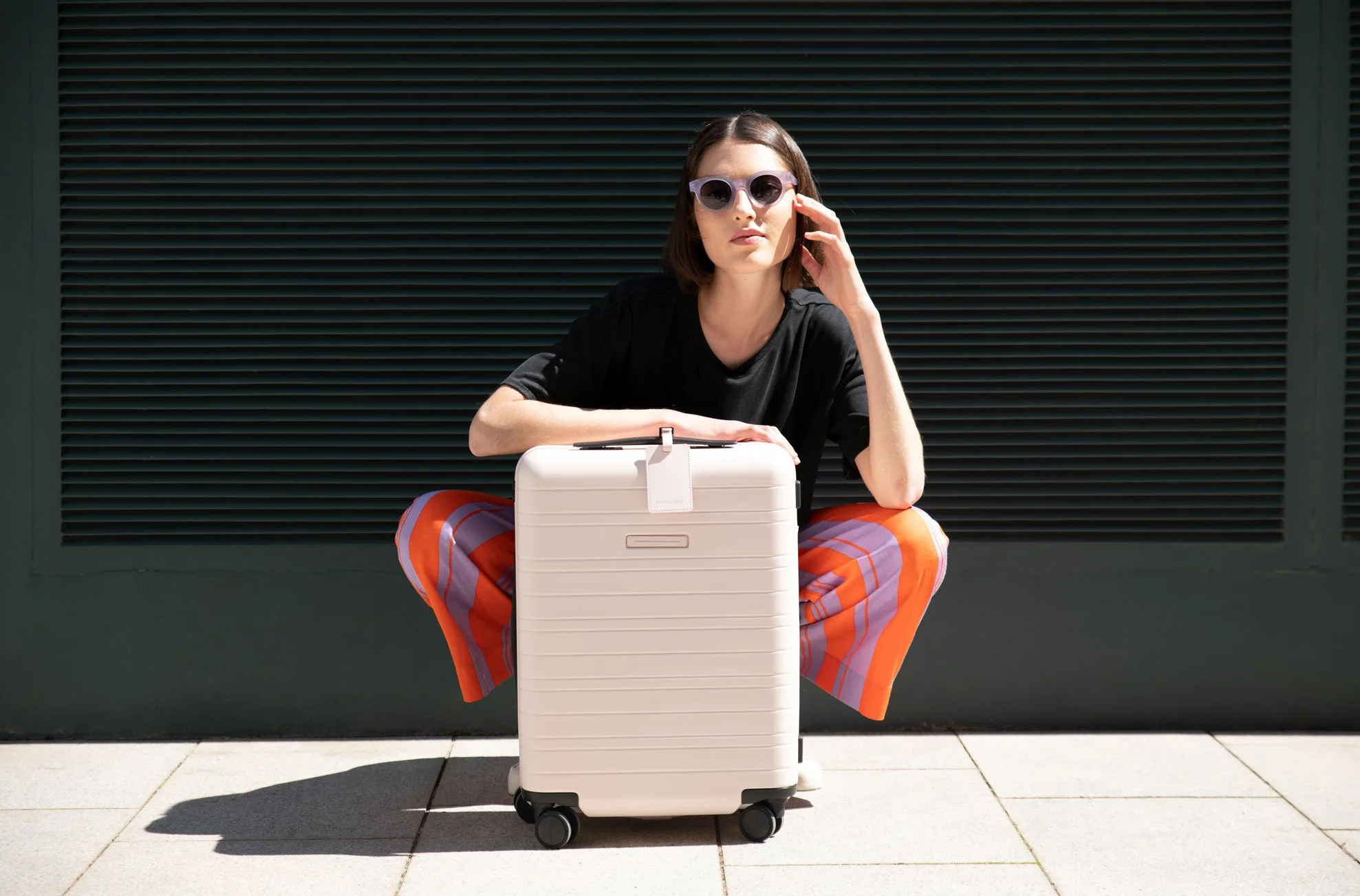 Woman sitting on the ground with a large white suitcase in front of her, wearing sunglasses, a black shirt, and bright striped pants, against a dark wall.