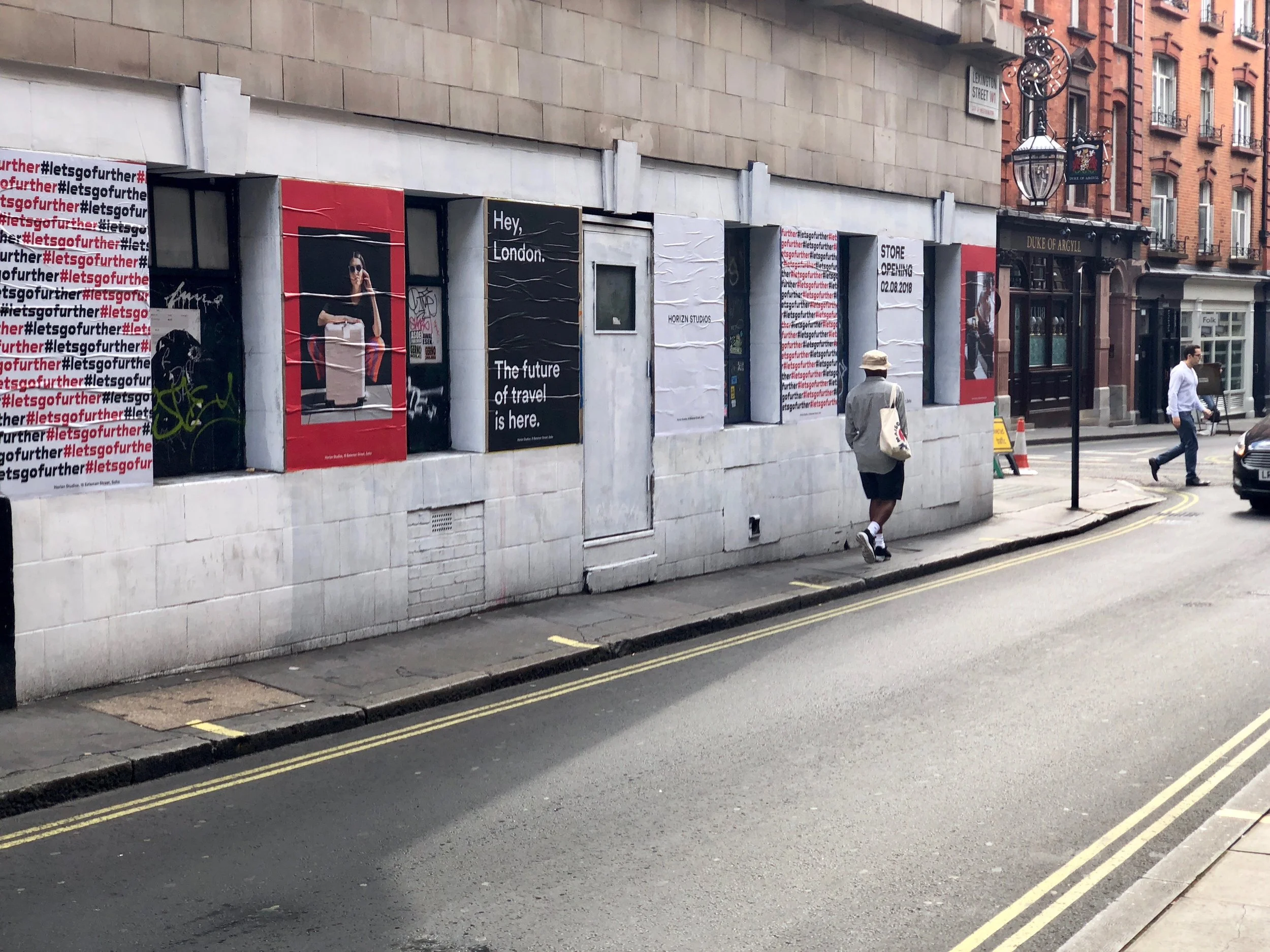A street scene in London with a sidewalk, a person walking, and a row of storefronts and advertisements on a building wall.