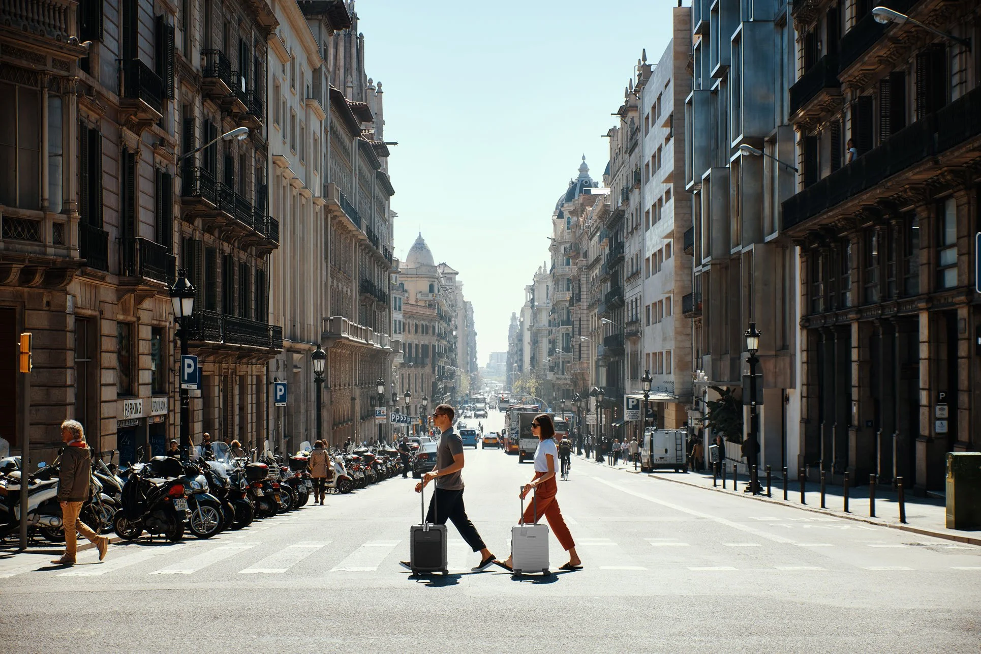 Two people crossing the street with rolling suitcases in a city with tall historic buildings and parked motorcycles on the side.