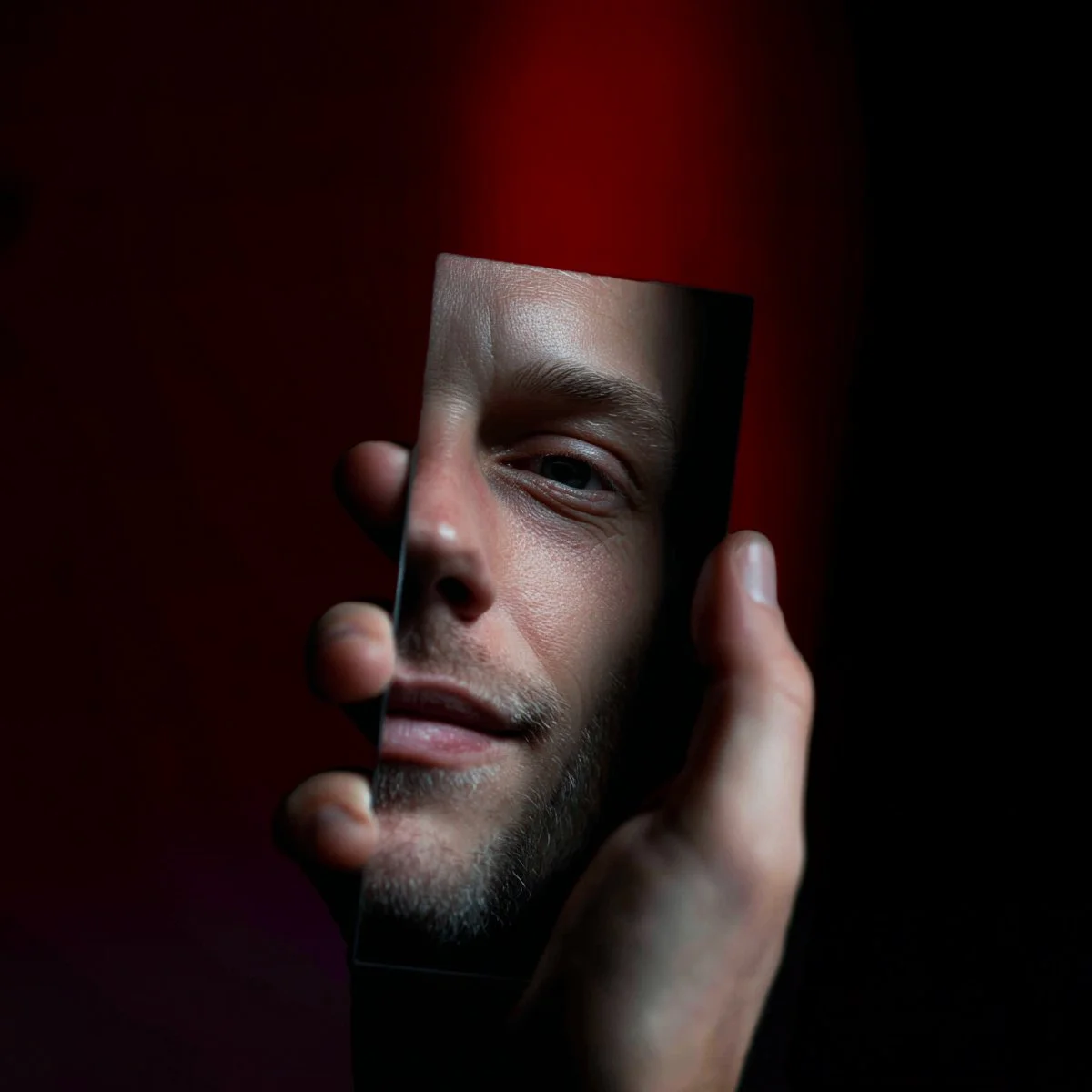Une main tient un miroir rectangulaire qui reflète le visage d'un homme avec une expression neutre, face sombre avec un fond noir et rouge derrière lui.