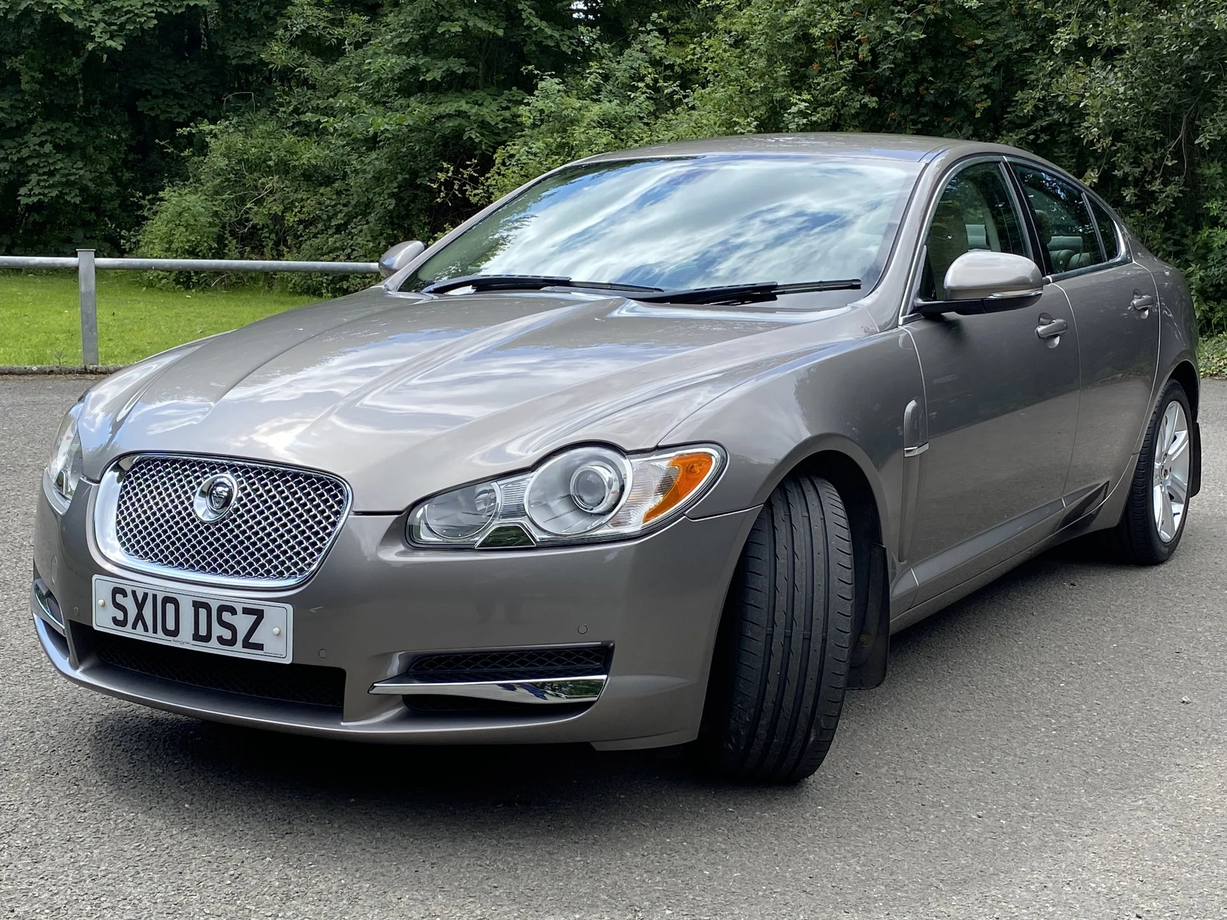 A silver Jaguar sedan parked on a gray asphalt surface with green trees and grass in the background.