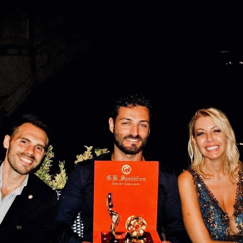 Tre persone sorridenti, uomini e donna, seduti con uno schermo rosso tra loro, in un evento serale. La donna indossa un vestito decorato con paillettes, gli uomini sono elegantemente vestiti.