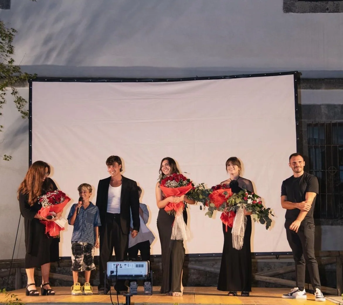 Gruppo di persone che festeggiano sul palco, alcune con bouquet di fiori, sullo sfondo di un grande schermo bianco.