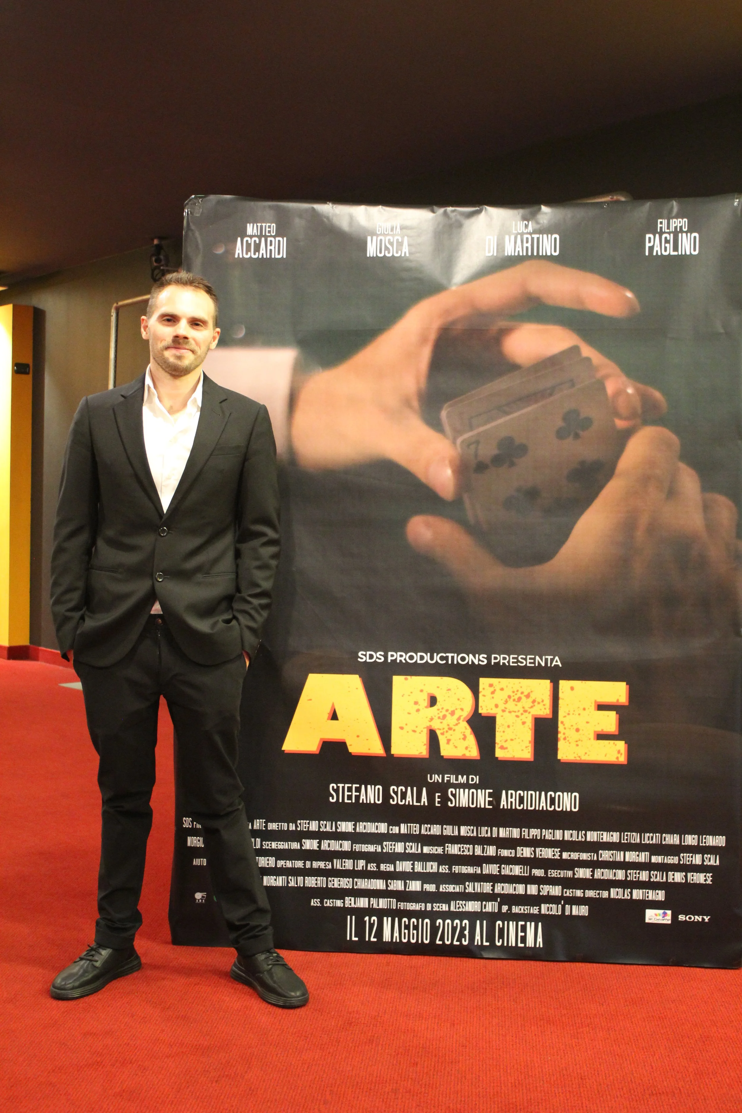 Un uomo vestito in completo nero si trova accanto a un poster del film "Arte" con i nomi degli attori e la data di uscita 12 maggio 2023.