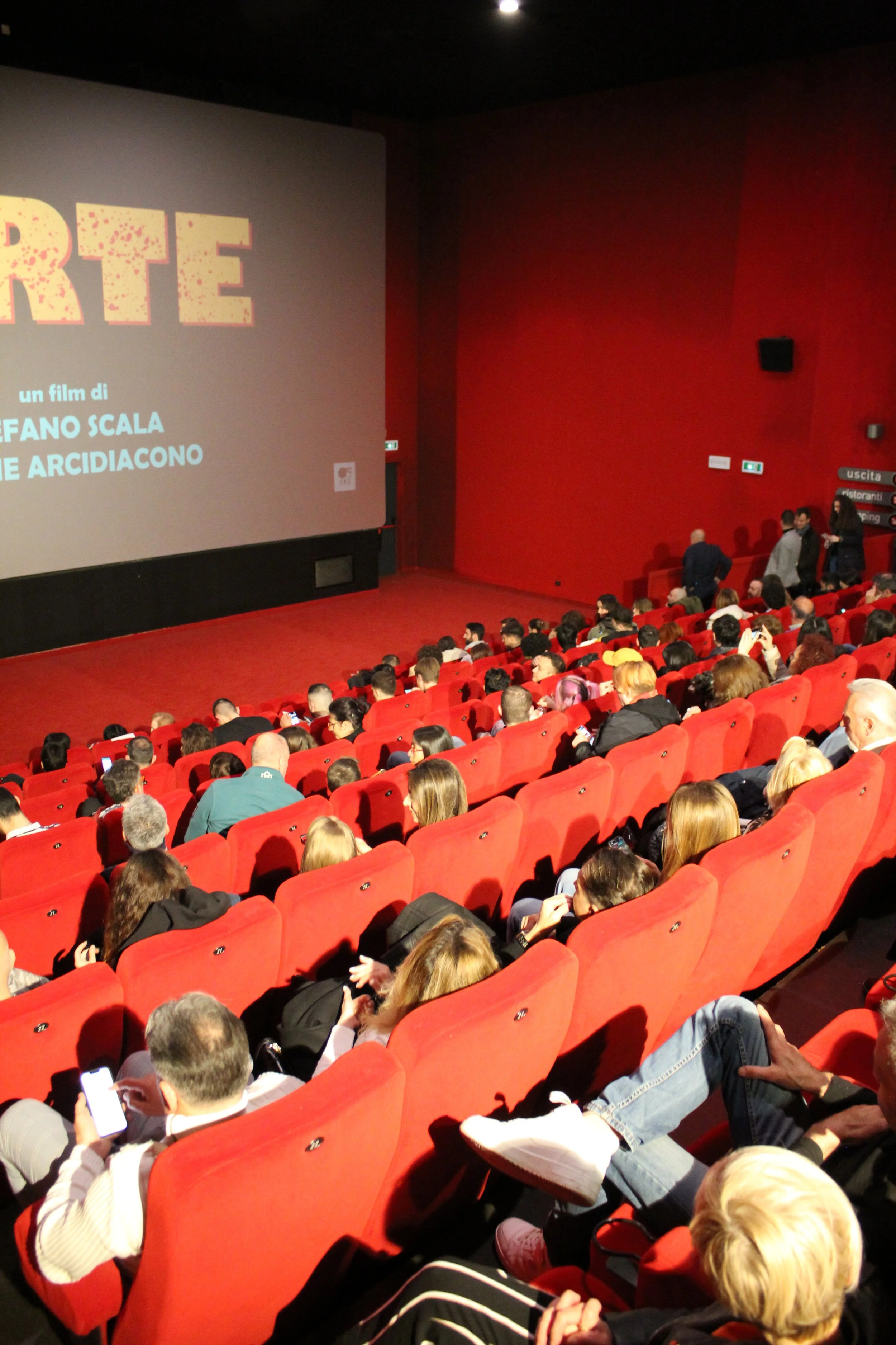 Interno di un cinema con persone sedute in poltrone rosse che guardano il grande schermo