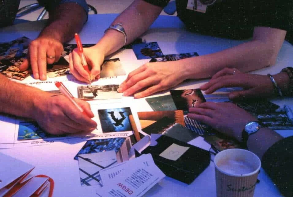 Multiple people working together, filling out photos and documents on a cluttered table at a meeting or event.