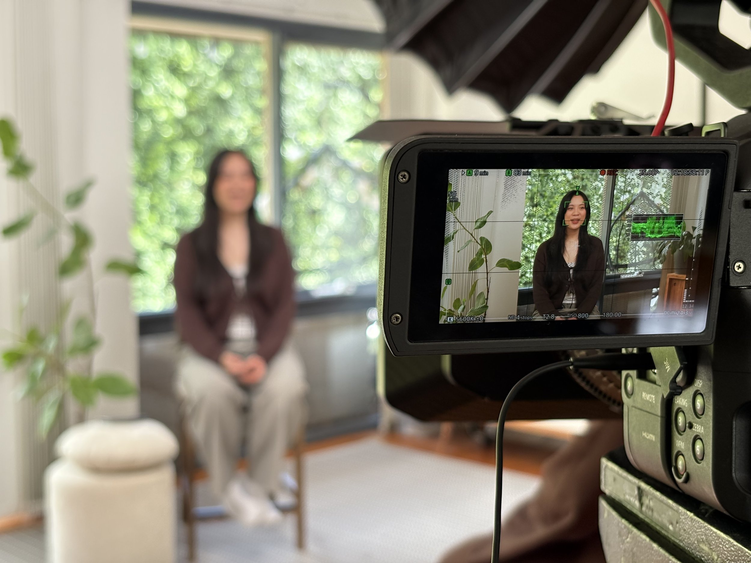A woman sitting on a chair indoors during a video recording, with a camera recording her in front of a large window and green plants.