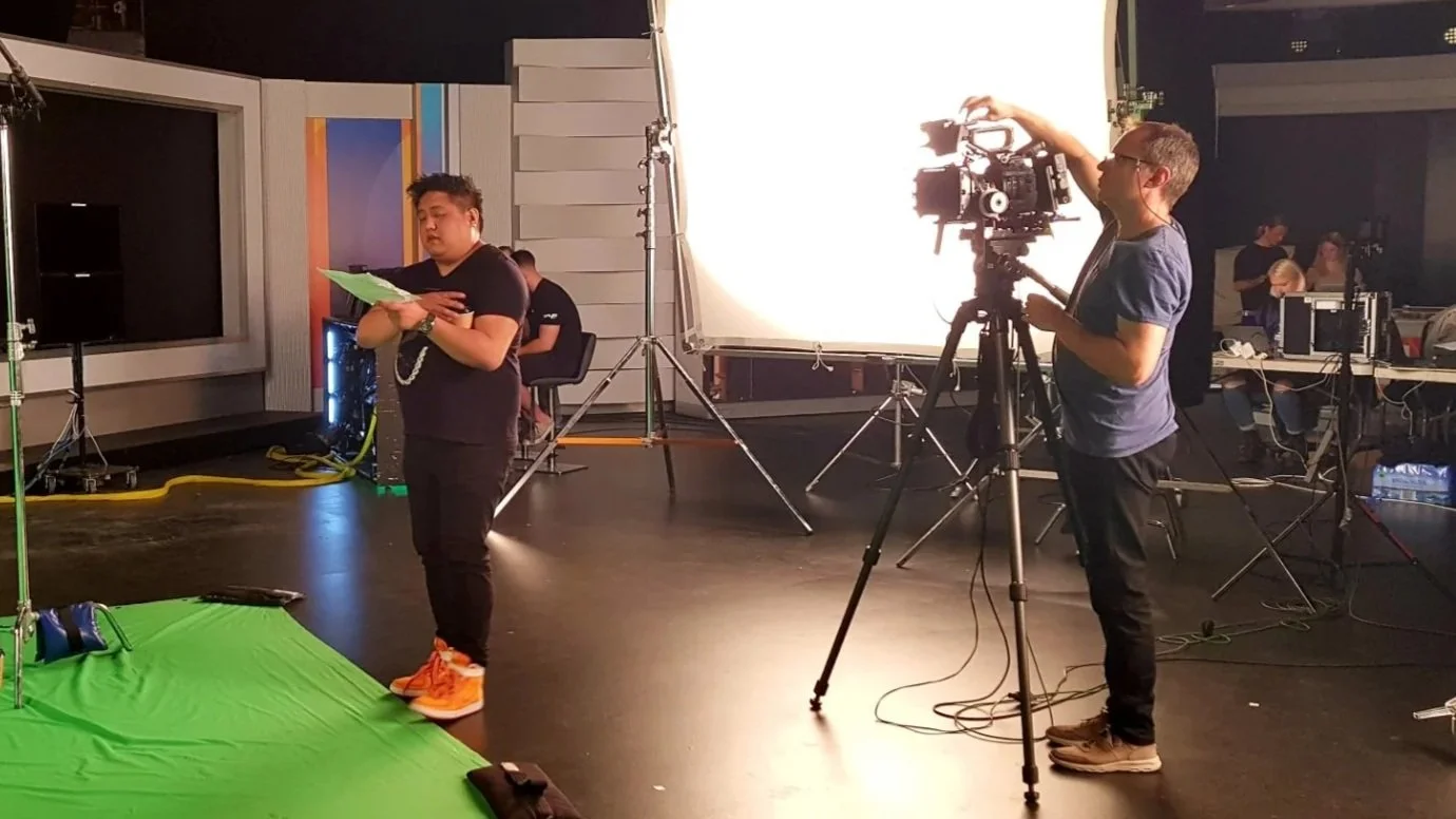 Behind the scenes of a video shoot with a green screen, a man holding a script, and a camera operator adjusting a large professional camera.