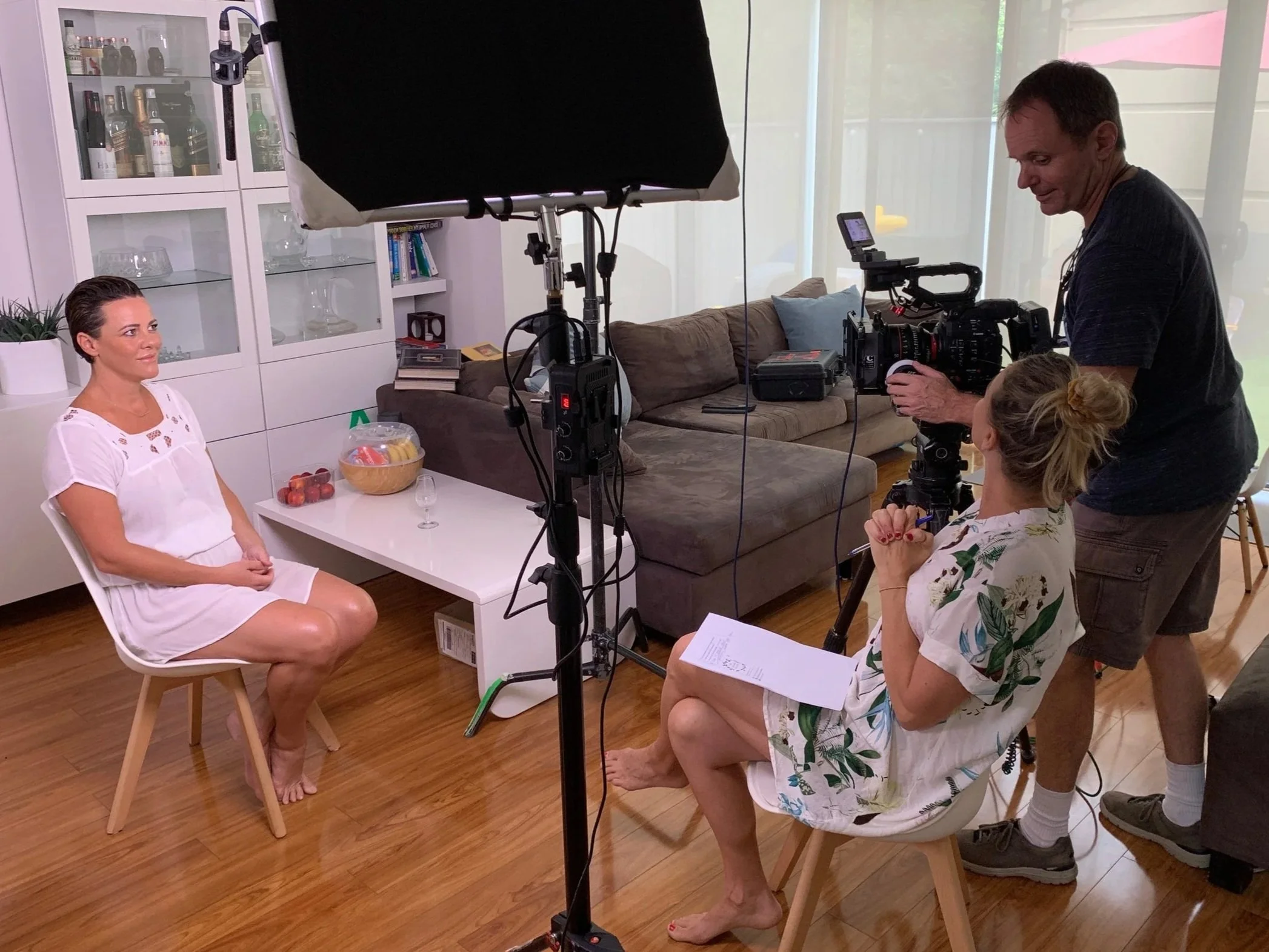 Filming a woman in a white dress for an interview or video shoot in a living room with camera crew and lighting equipment.
