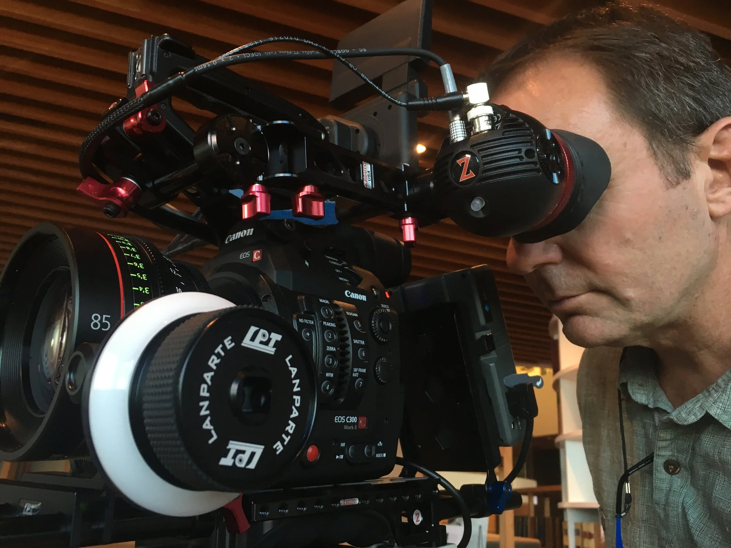 A man operating a professional video camera with a large lens and mounted accessories, indoors with wooden ceiling panels in the background.