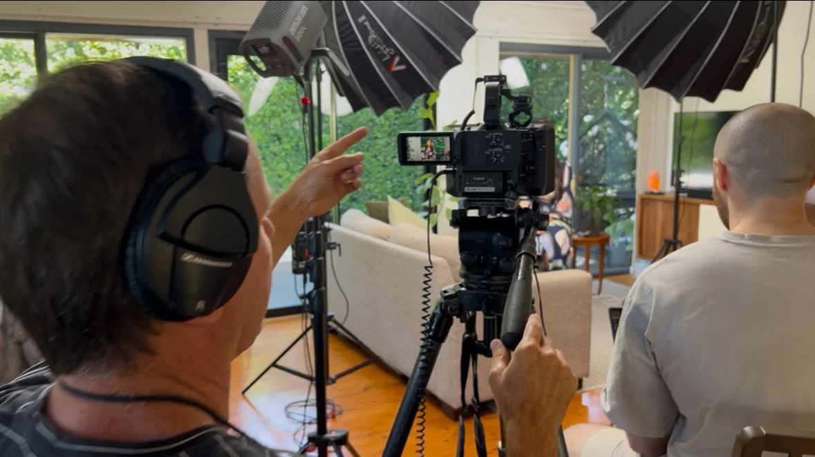 Filmmaking setup with a camera, lighting, and a crew member pointing at a shot in a living room with large windows and green foliage outside.