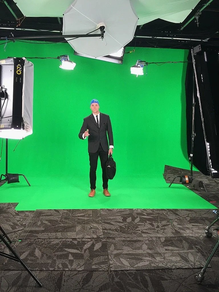 Man in a black suit, white shirt, and black tie standing in front of a green screen with studio lighting, holding a black bag.