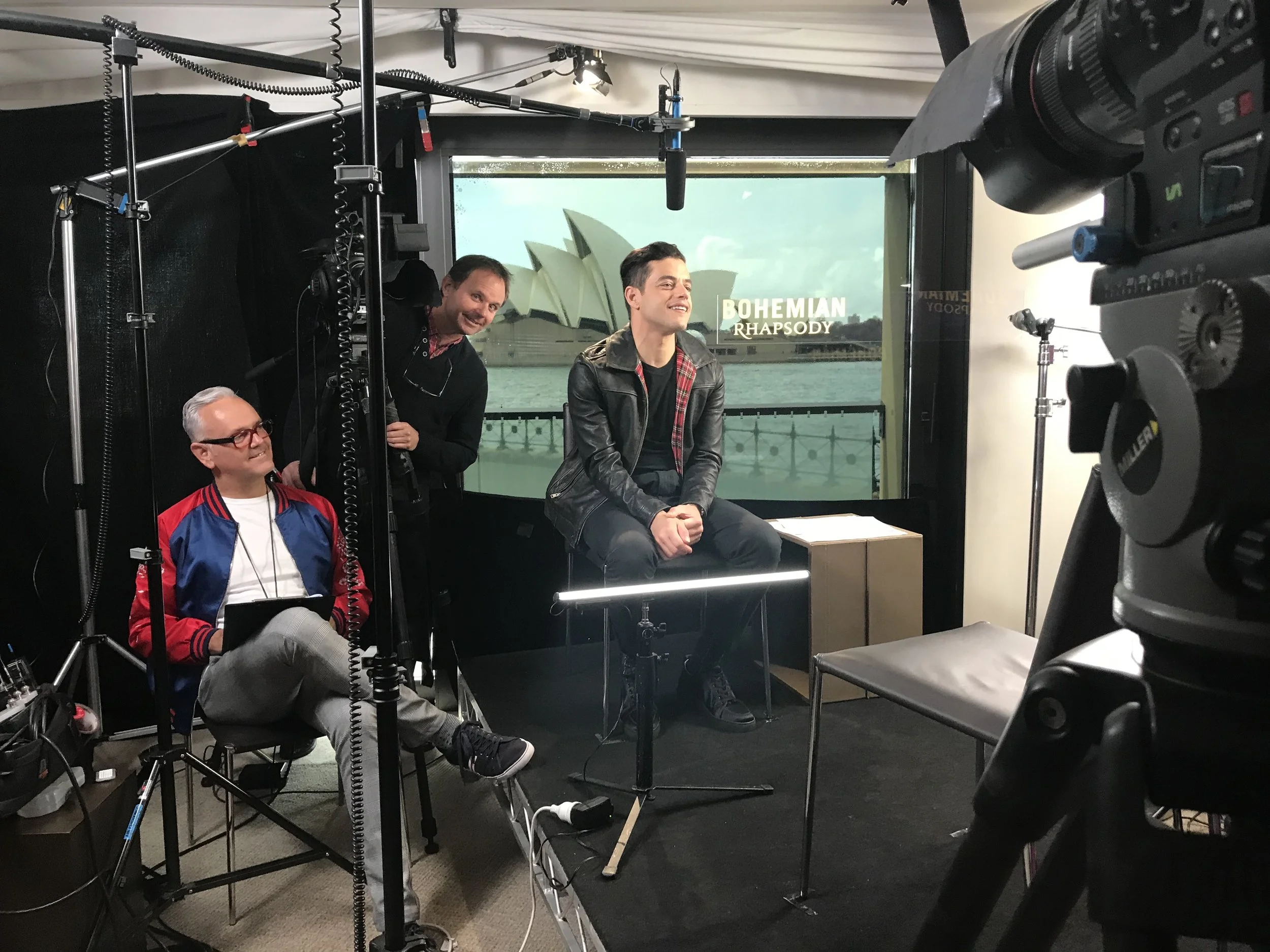 A filming studio with two men and one woman, one man sitting on a stool, the woman and the other man standing nearby, large camera in the foreground, webcam on the window in the background, and a view of the Sydney Opera House