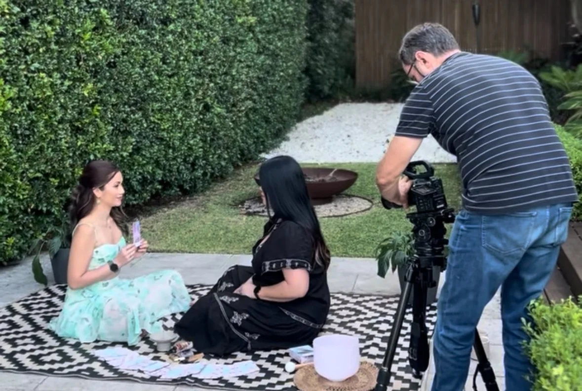A woman sitting on a black and white patterned rug on a patio, holding playing cards, with another woman sitting across from her and a man filming them with a professional camera.