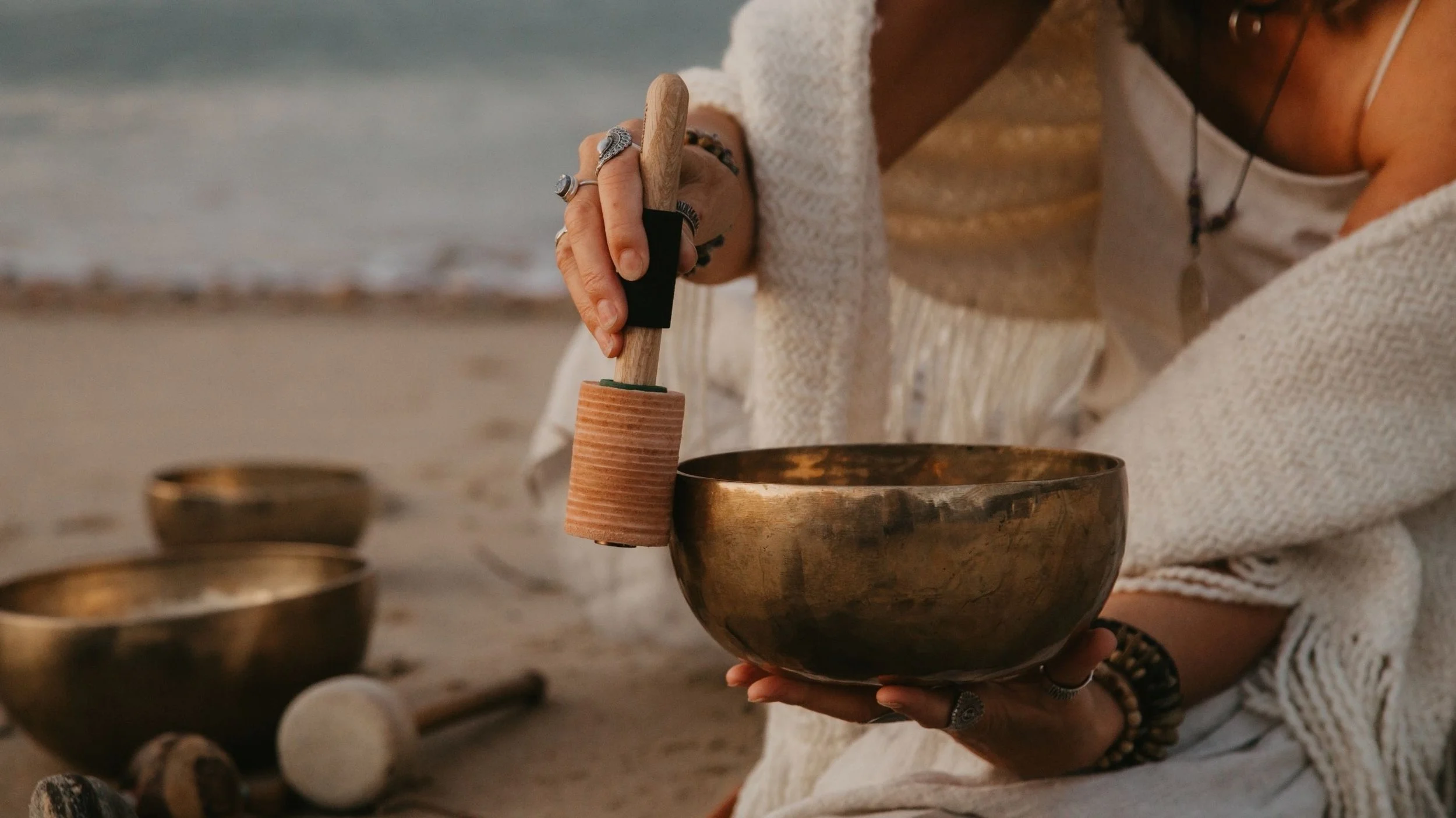 Close up of a sound practitioner holding a bronze, handmade Himalayan singing bowl. She is striking the bowl on the rim. In the background is the sea