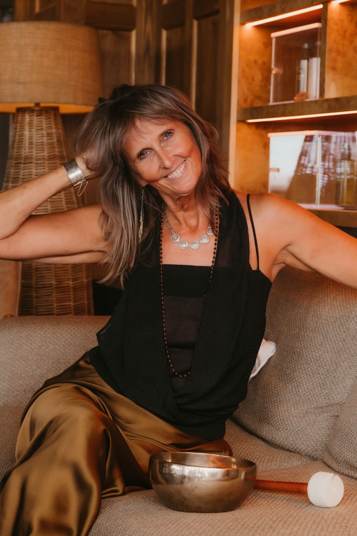 Alison Palmer Sound Practitioner sits on a couch with a golden singing bowl by her hip. Shis is wearing smart clothes and looks like a professional sound healer