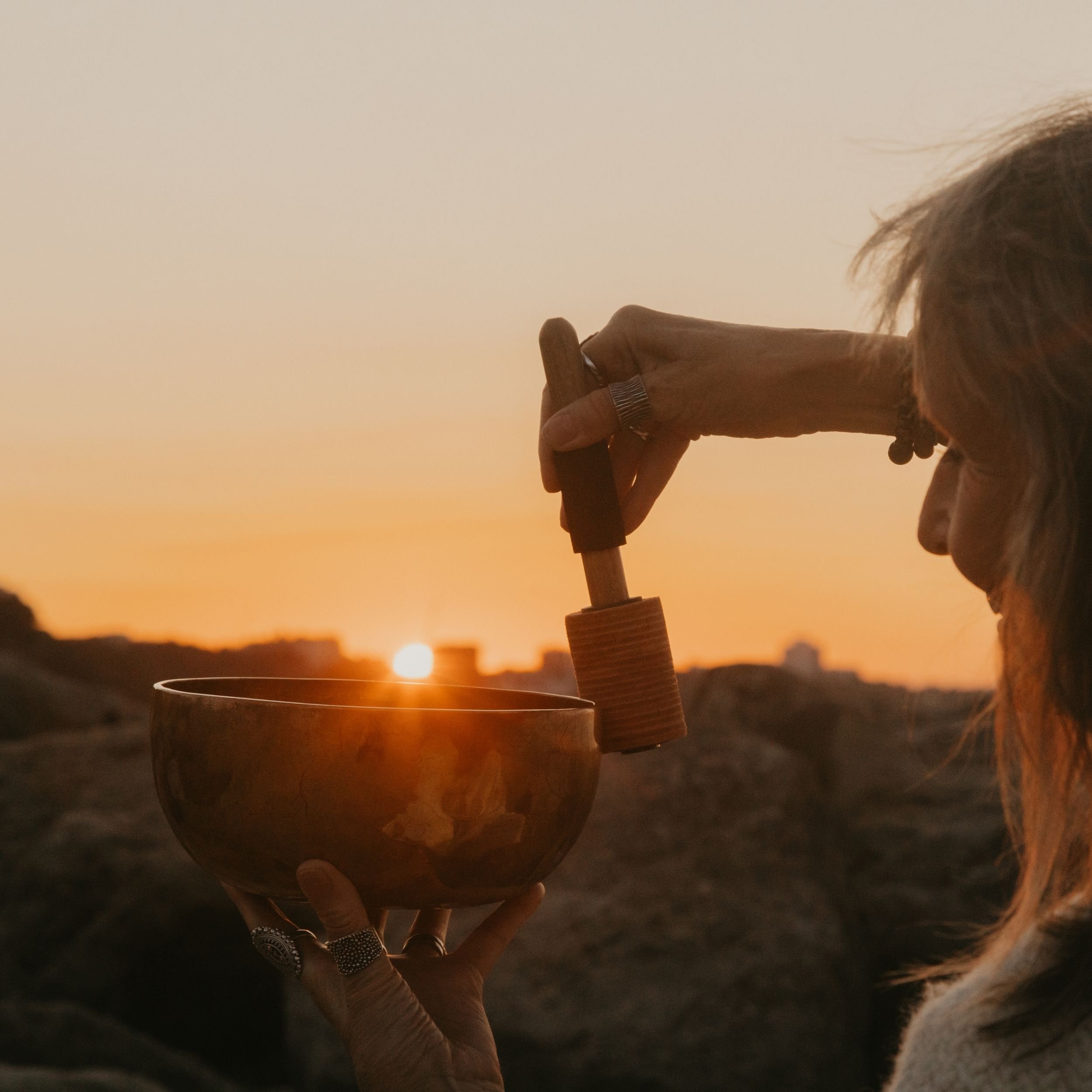 A Sound Healing Teacher demonstrates playing a bronze Himalayan Singing Bowl outside at sunrise. The sun is rising and the angle of the camera makes it appear to emerge from the bowl