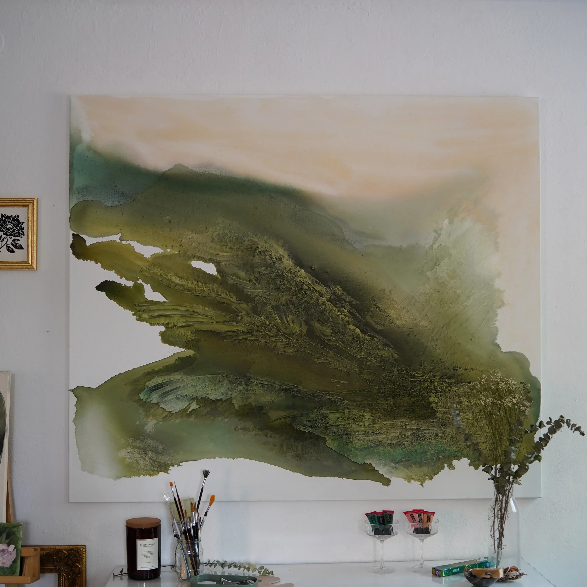 Abstraktes Wandbild in Grüntönen an einer weißen Wand, daneben kleine Dekorationsgegenstände und ein Vase mit Zweigen.