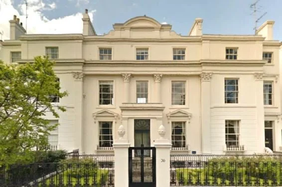 A large white historic building with multiple windows, a central door, and decorative architectural details, enclosed by a black gate and surrounded by greenery.