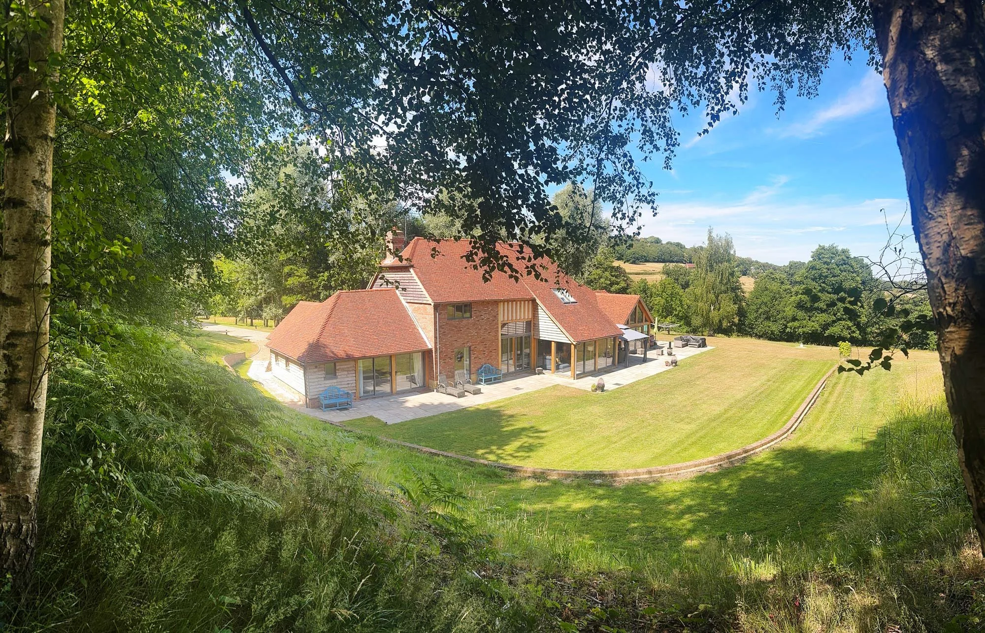 Charming red-brick cottage set on expansive clipped lawn, framed by mature oak trees and countryside backdrop.