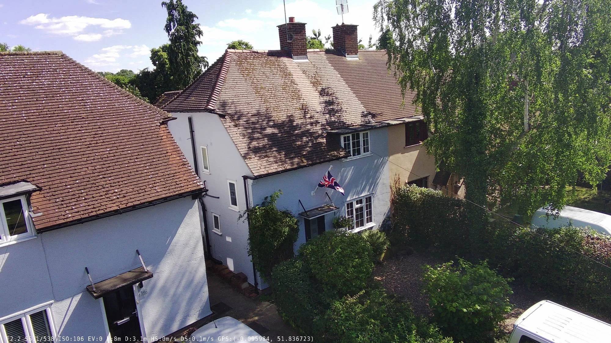 Row of narrow terraced English cottages with hanging flower baskets and mature trees, seen from above.