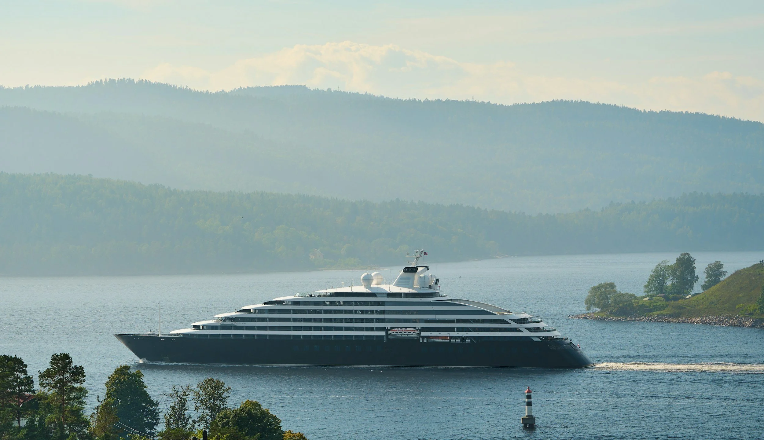 Large luxurious yacht sailing on calm water with forested hills and mountains in the background.