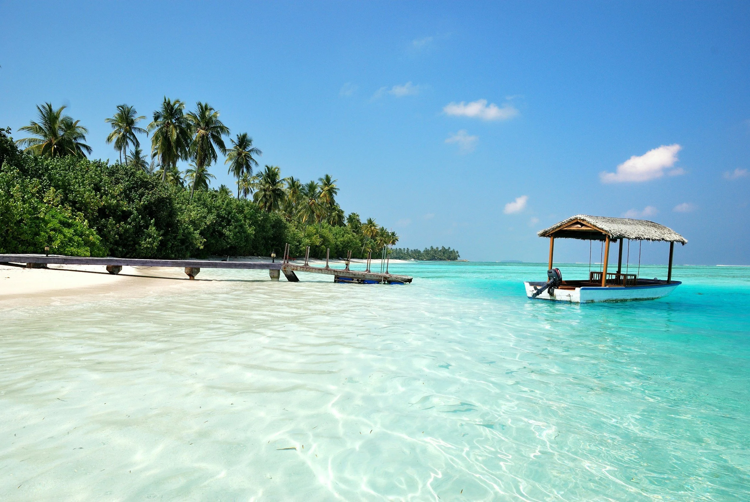 Tropical beach with white sand, lush green trees, a wooden dock, a small boat with a thatched roof, and clear turquoise water under a blue sky with a few clouds.