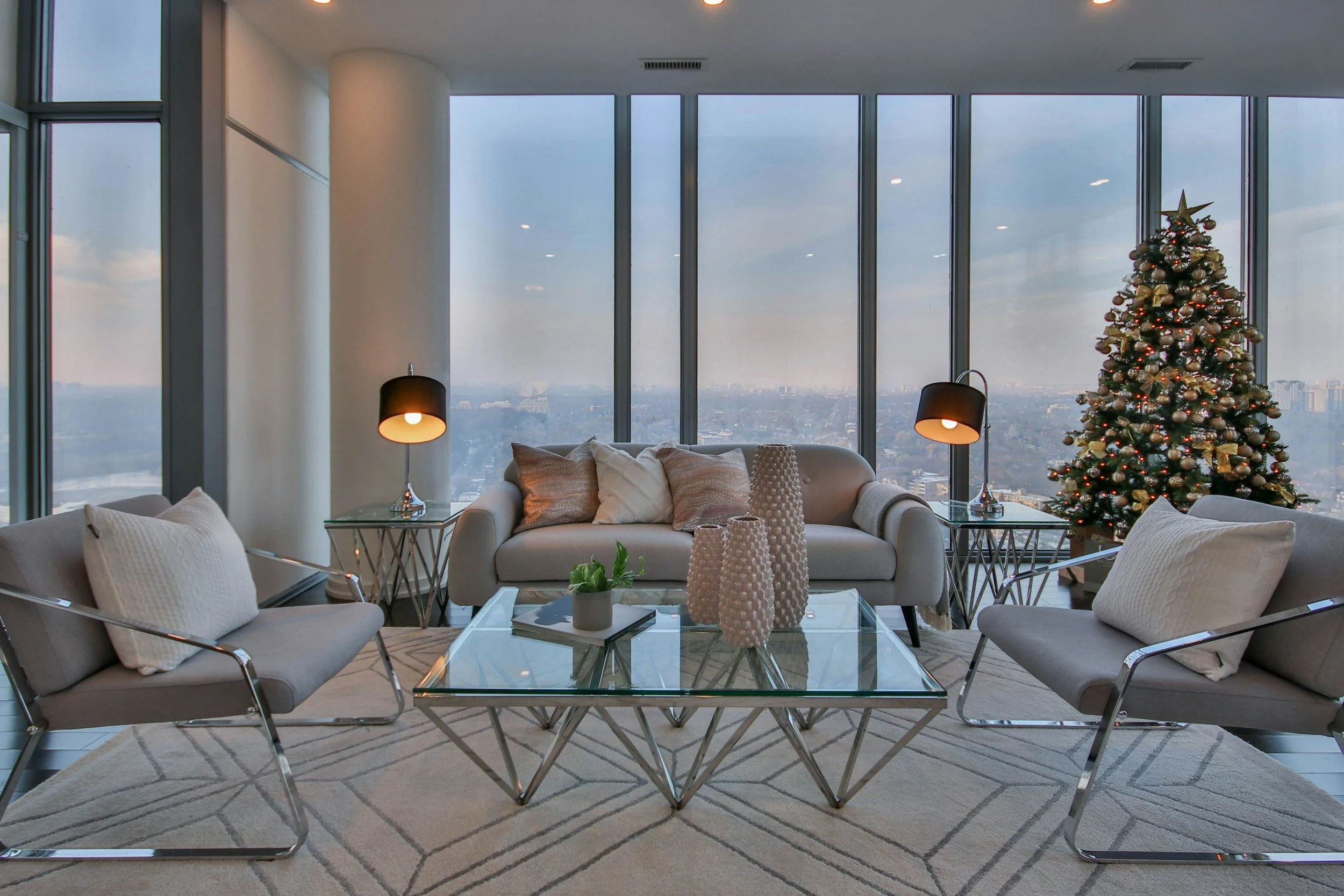 Modern Luxury hotel suite with large windows, a decorated Christmas tree, white sofas with pillows, glass coffee table, and black lamps, overlooking a cityscape.