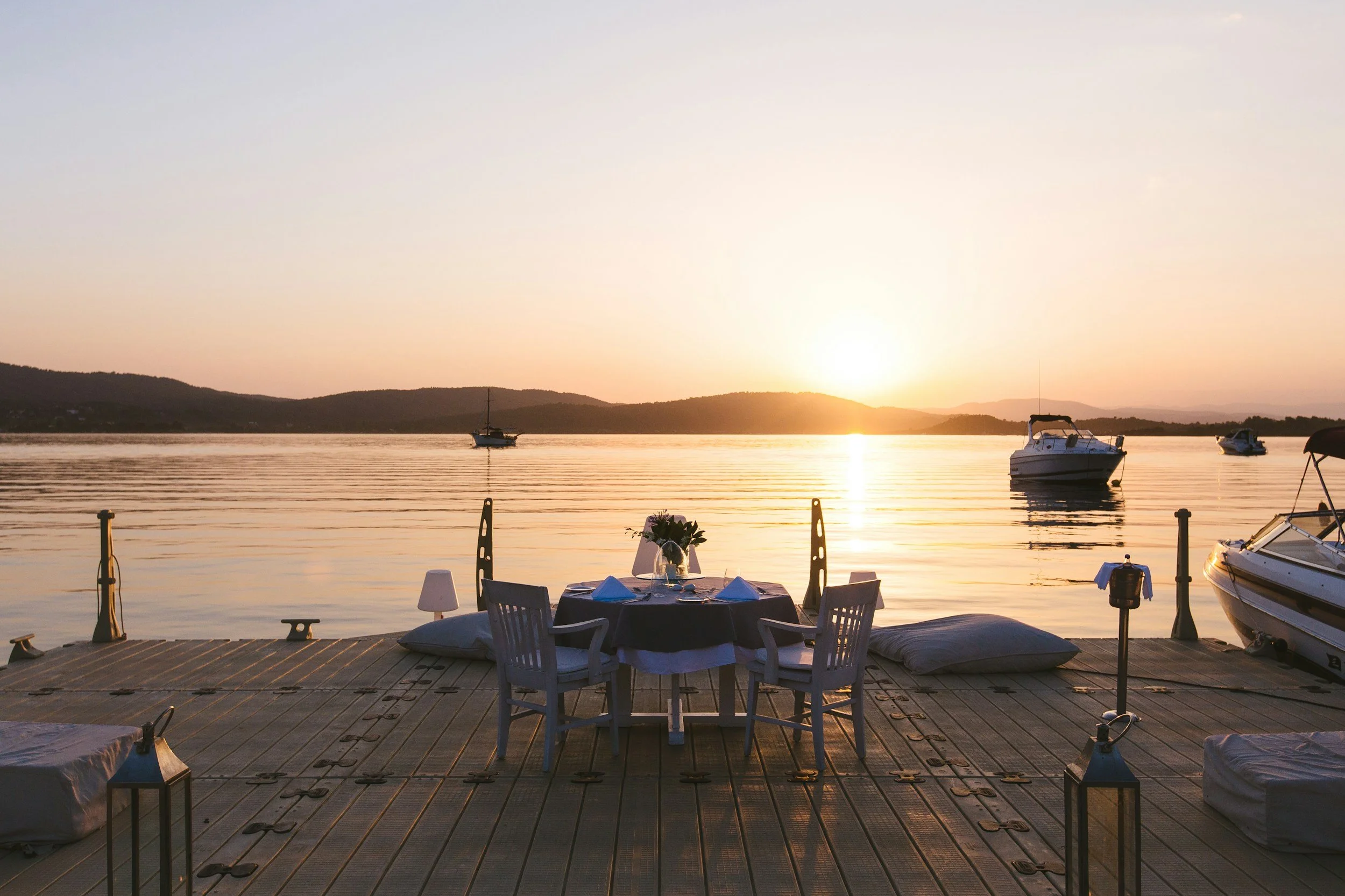 A lakeside view during sunset with boats on the water and a table set for dining on a wooden dock.