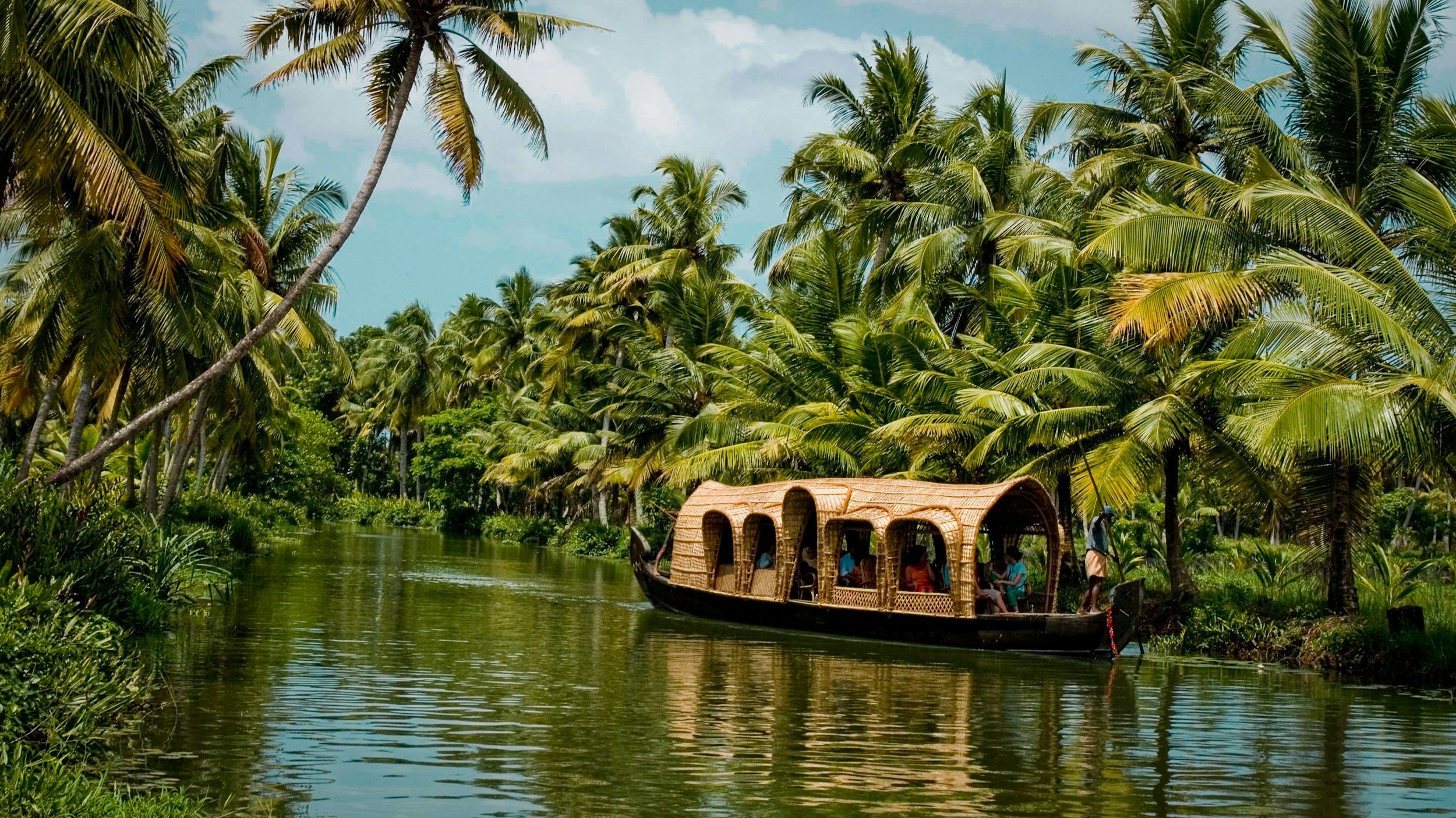 A boat with a bamboo-covered roof carrying people on a river surrounded by lush green palm trees.