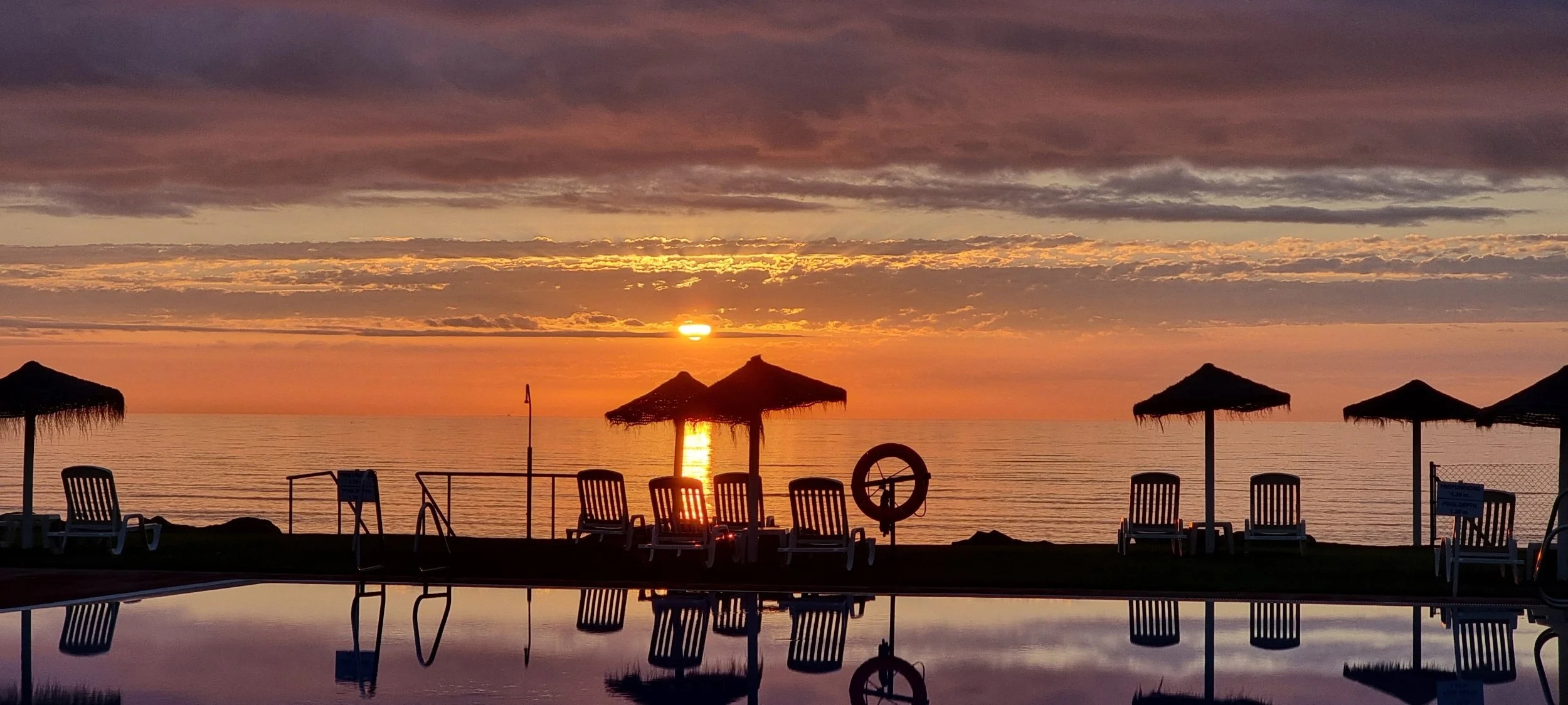 Sunset over the ocean with beach umbrellas and lounge chairs near the water, reflected in a pool in the foreground.