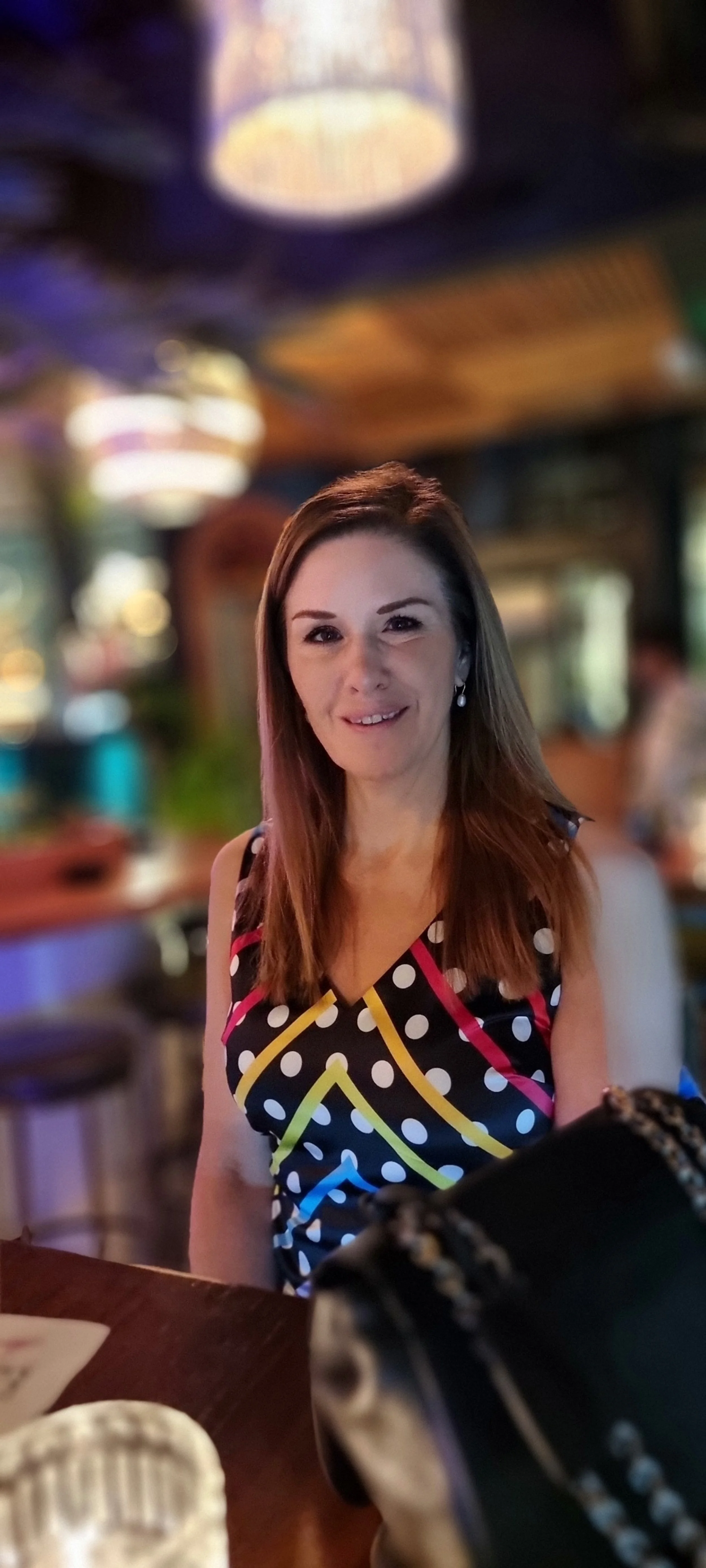 A woman with long brown hair, wearing a sleeveless black dress with colorful polka dots and geometric patterns, sitting at a wooden table in a dimly lit restaurant or bar with blurred background. She is smiling slightly and looking at the camera.