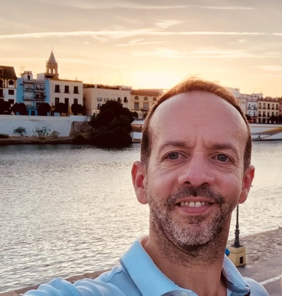 A man taking a selfie near a body of water during sunset, with buildings and a church steeple in the background.