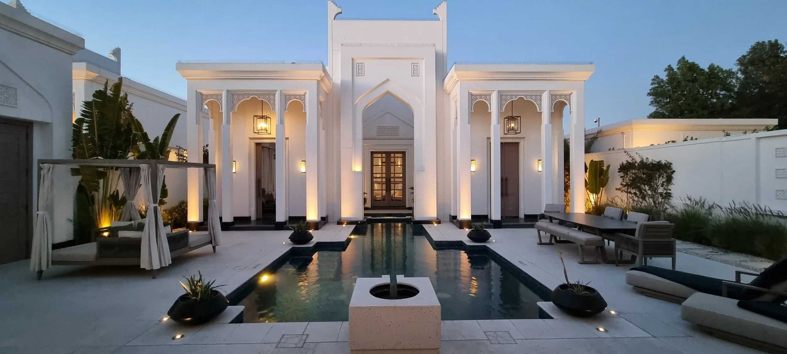 An elegant modern house with white walls, a small water pool in the center, outdoor seating, and decorative plants, illuminated by soft lighting at dusk.