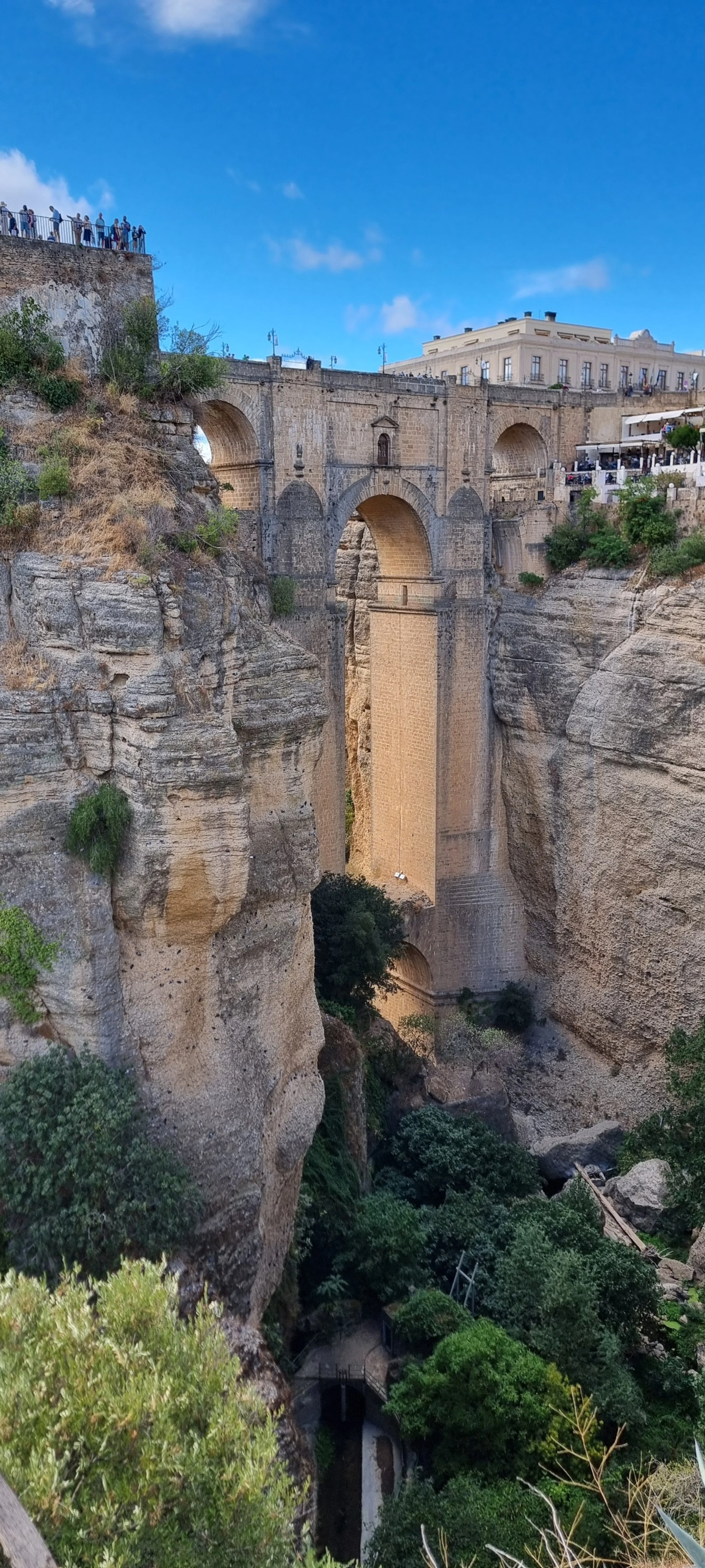 Historic stone bridge with multiple arches spanning a deep canyon with lush greenery and rocky cliffs, against a bright blue sky.