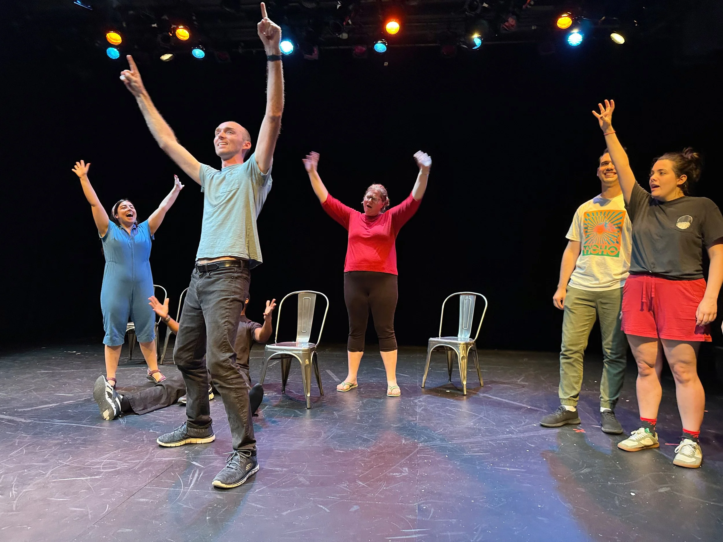 Group of six people on stage participating in an improv or theater activity, some standing with arms raised, others seated or kneeling, with chairs behind them, stage lights overhead, dark background.