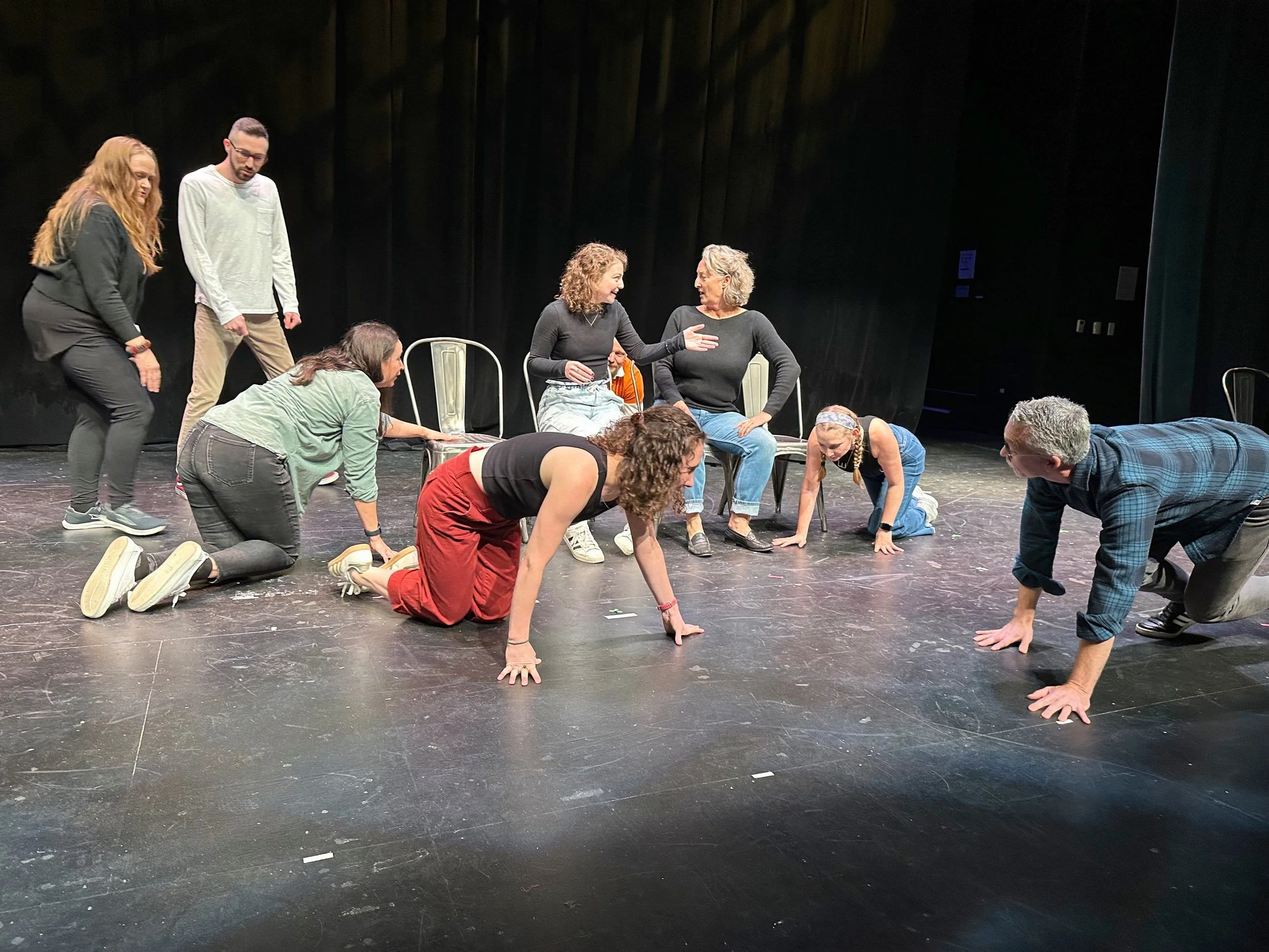 A group of people participating in a theatrical rehearsal on a stage, some kneeling and others sitting or standing, engaged in a scene or discussion.