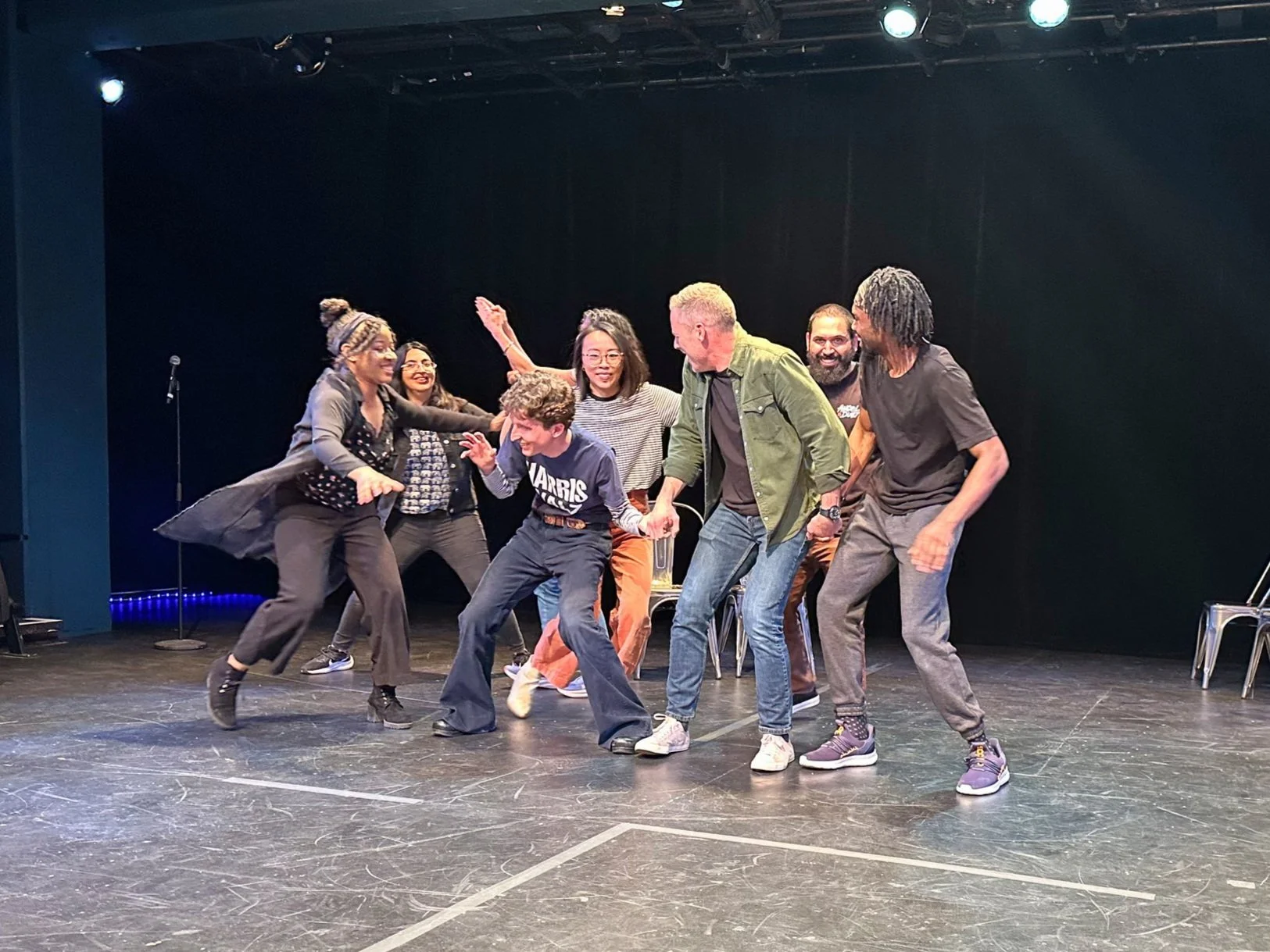 Group of people on stage dancing and laughing together, with a black background and stage lighting.