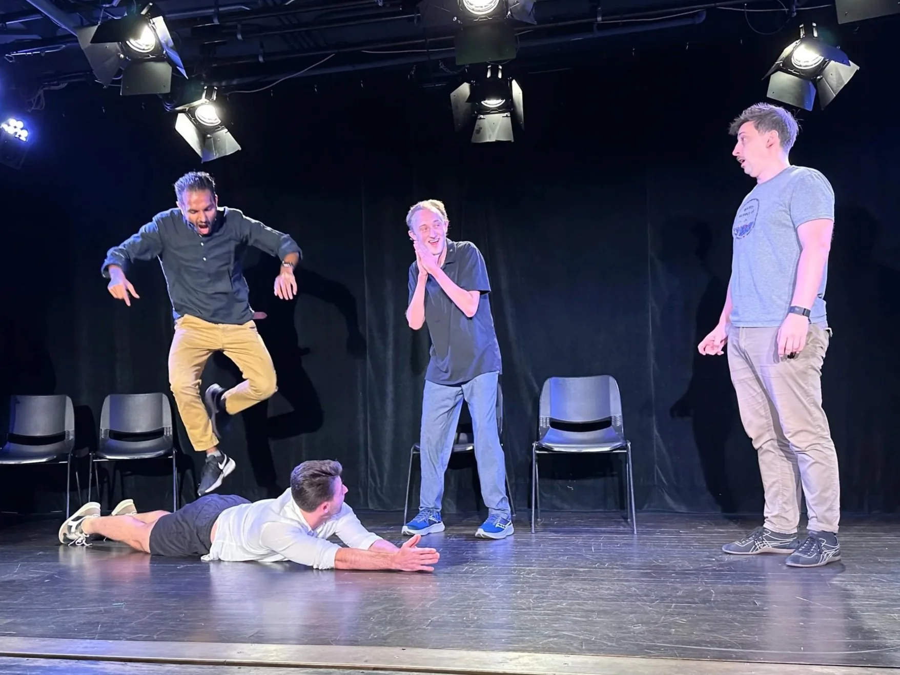 Four men performing a comedy skit or improv on stage, with one lying on the floor, one jumping in the air, and the others displaying surprised and amused expressions.