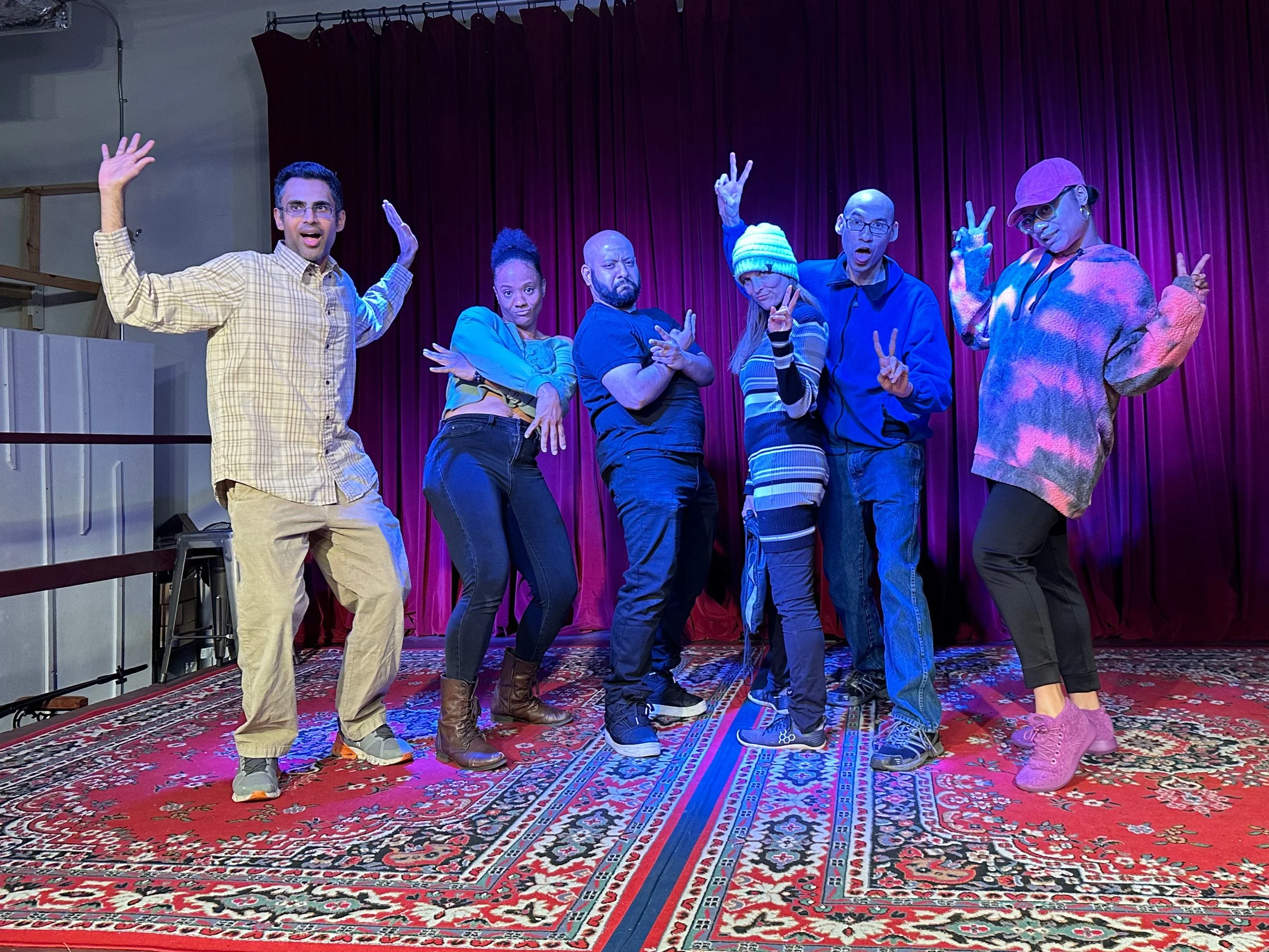 Six people standing on a stage with a red curtain, posing with playful gestures, under colorful stage lighting.
