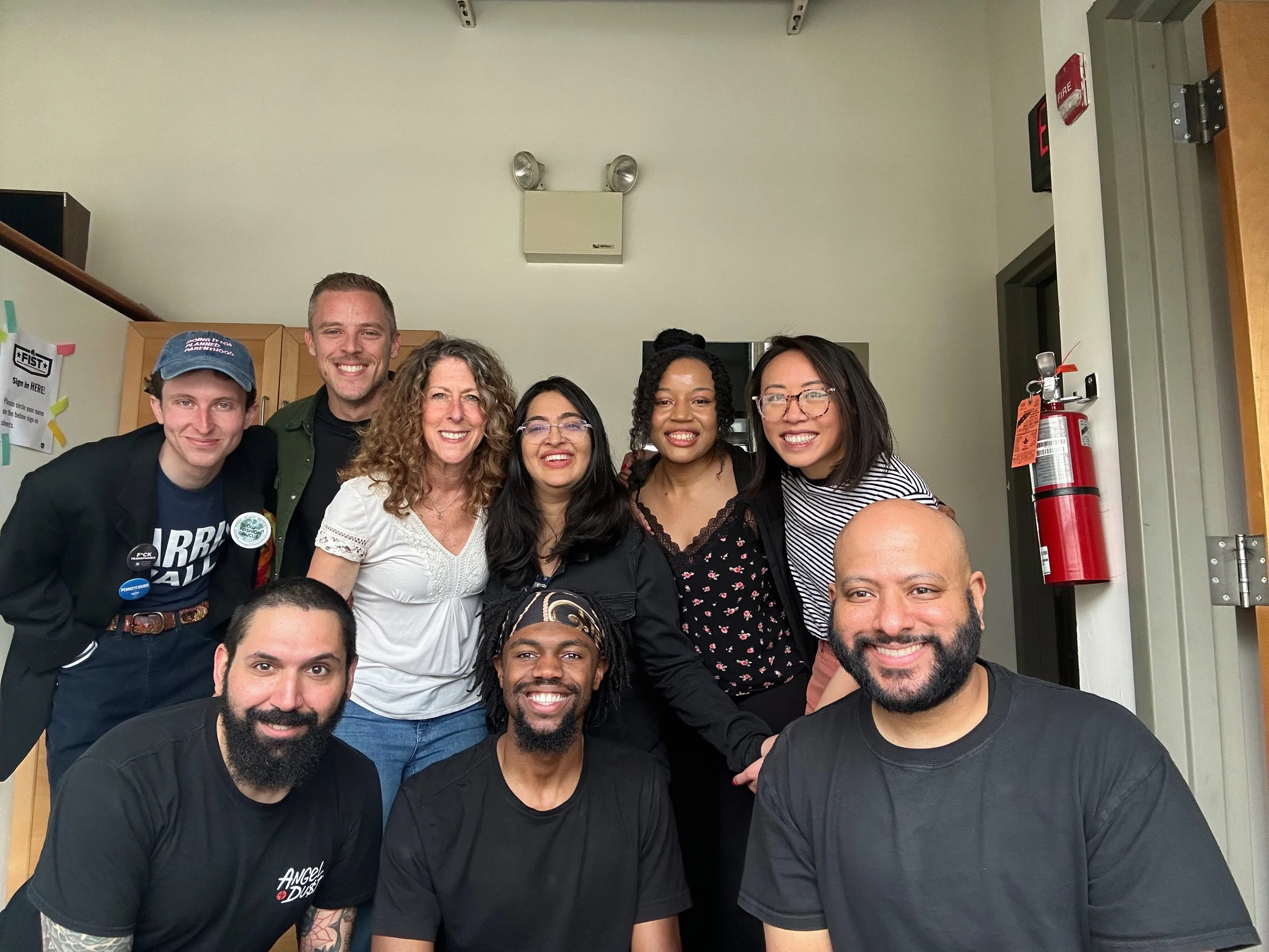 Group of ten diverse people smiling and posing for a photo indoors, with a whiteboard and a fire extinguisher in the background.