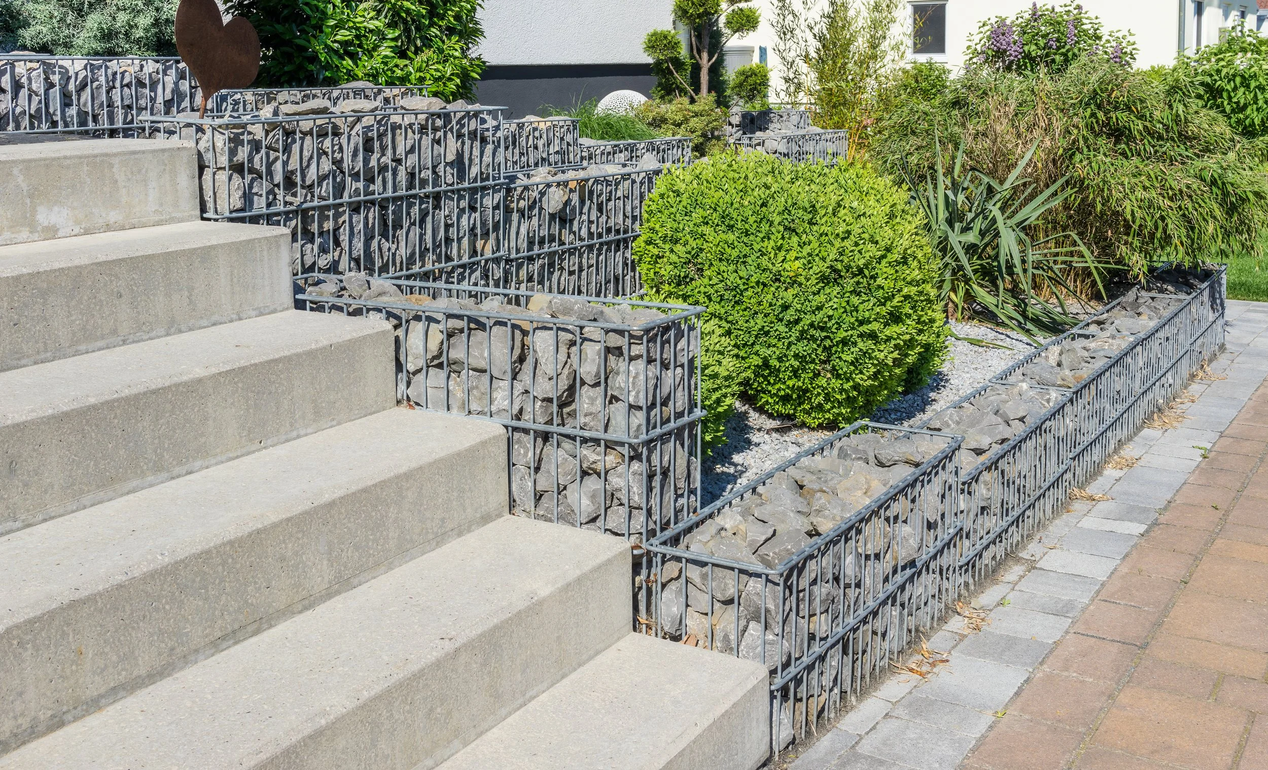 A modern outdoor space featuring gabion baskets filled with stones, used as retaining walls alongside concrete steps and lush green plants.