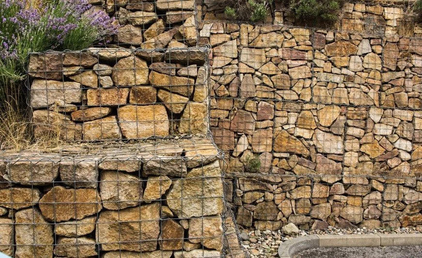 Gabion walls filled with stacked golden granite gabion stones, creating a sturdy retaining structure. The walls are surrounded by a few purple flowers and gravel.