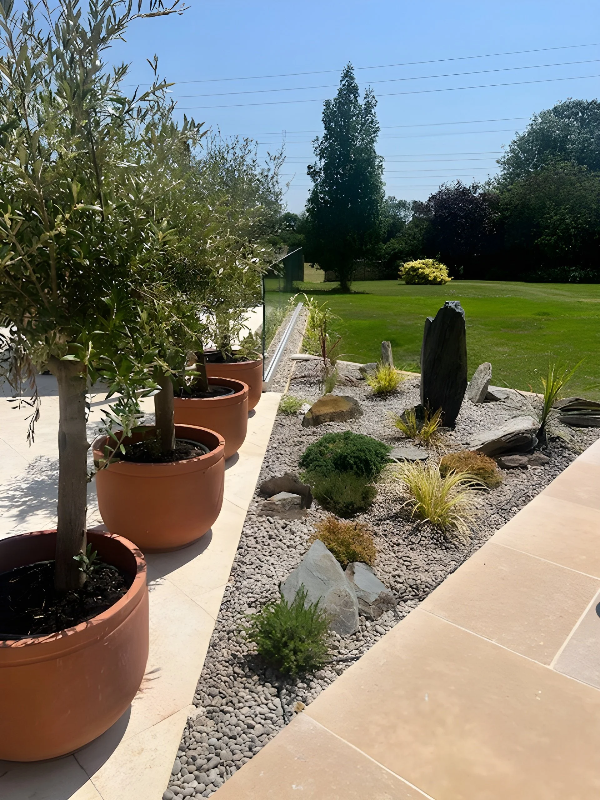 Alt text: A neatly landscaped garden area with potted olive trees along a paved walkway, featuring a rock and plant arrangement with gravel and decorative stones, set against a lush green lawn and trees in the background.