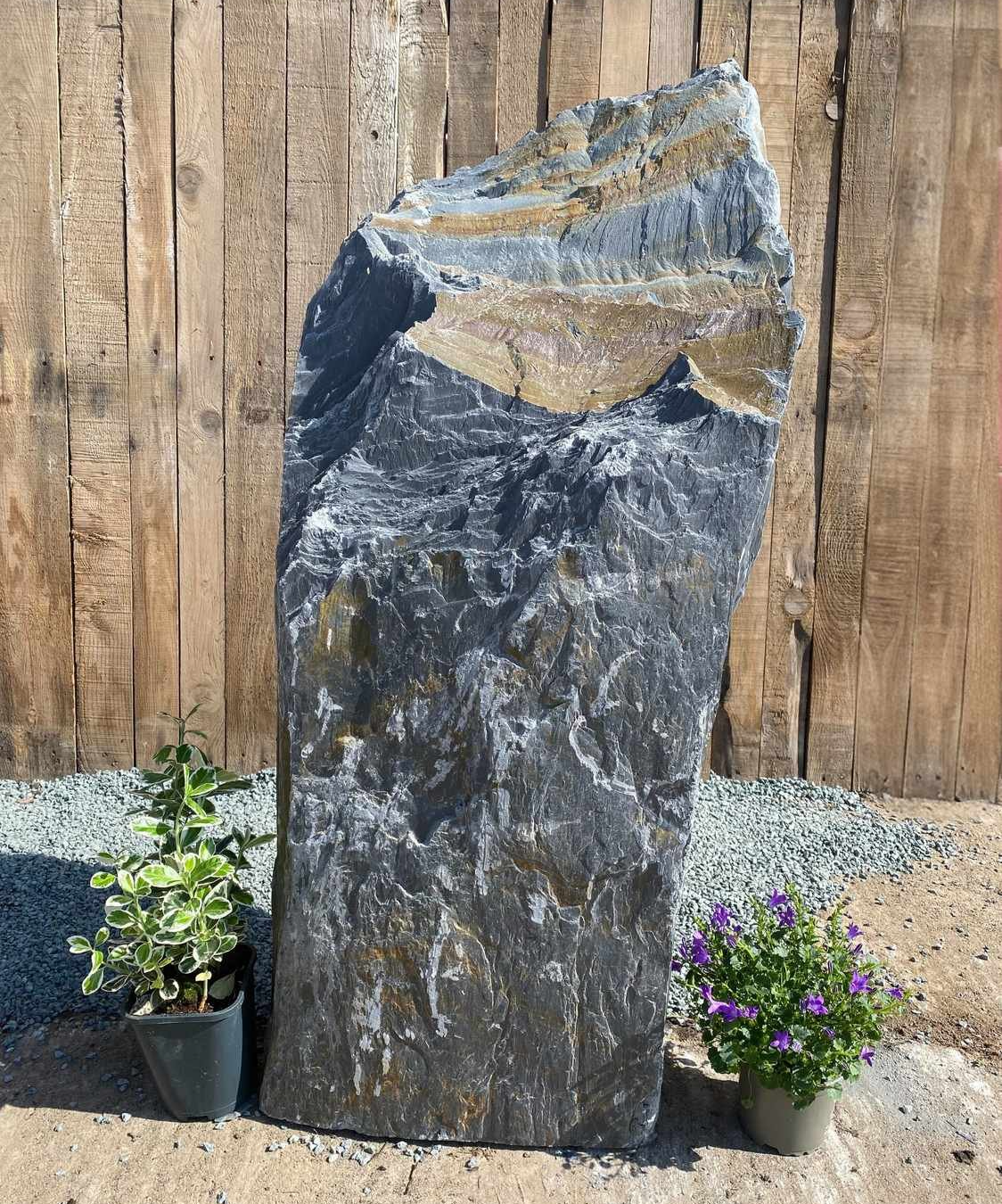 A large, textured monolith water feature that stands upright in a garden setting, positioned in front of a wooden fence.