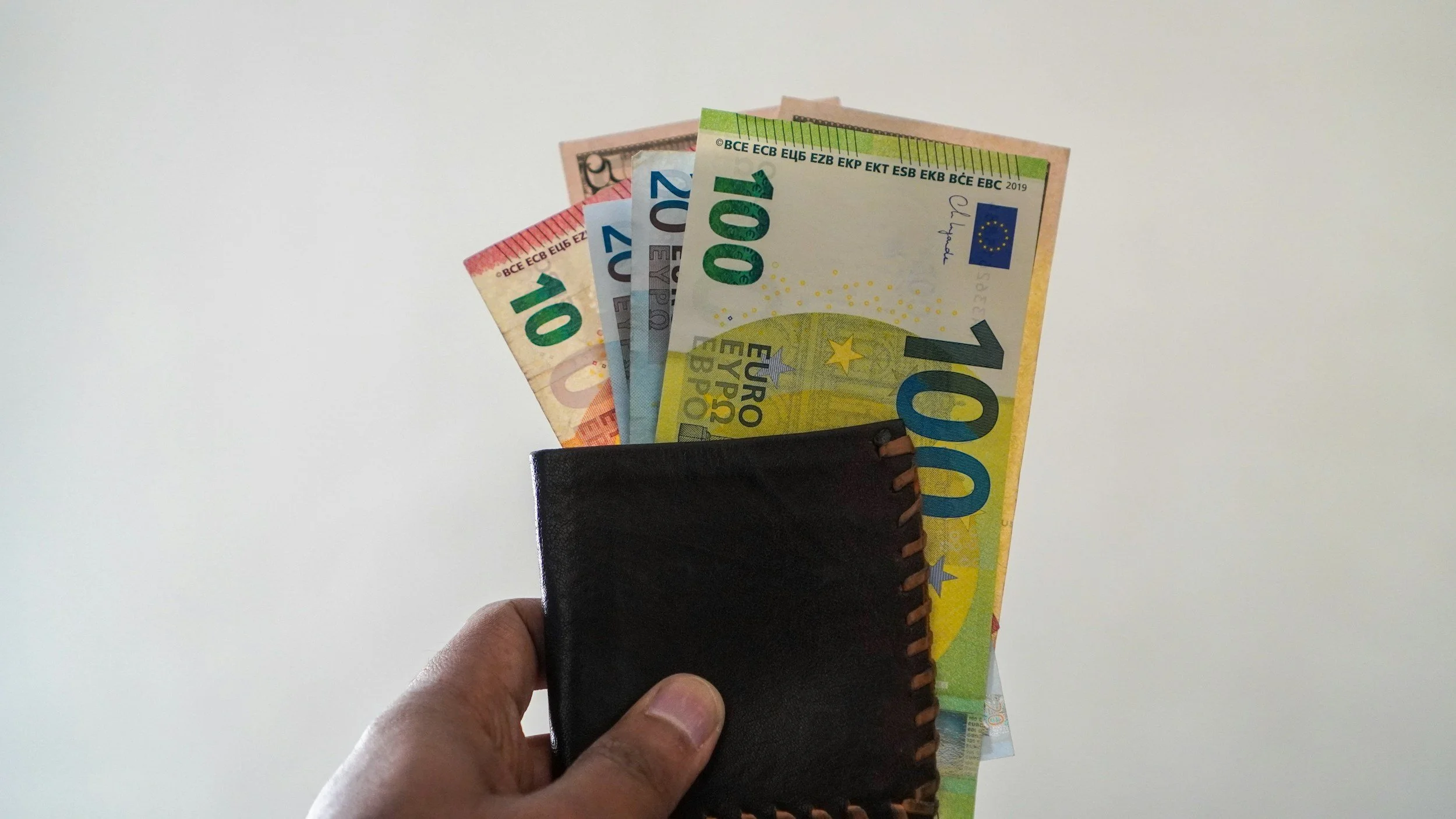 A hand holding a black leather wallet with four Euro banknotes partially extending from it against a plain background.