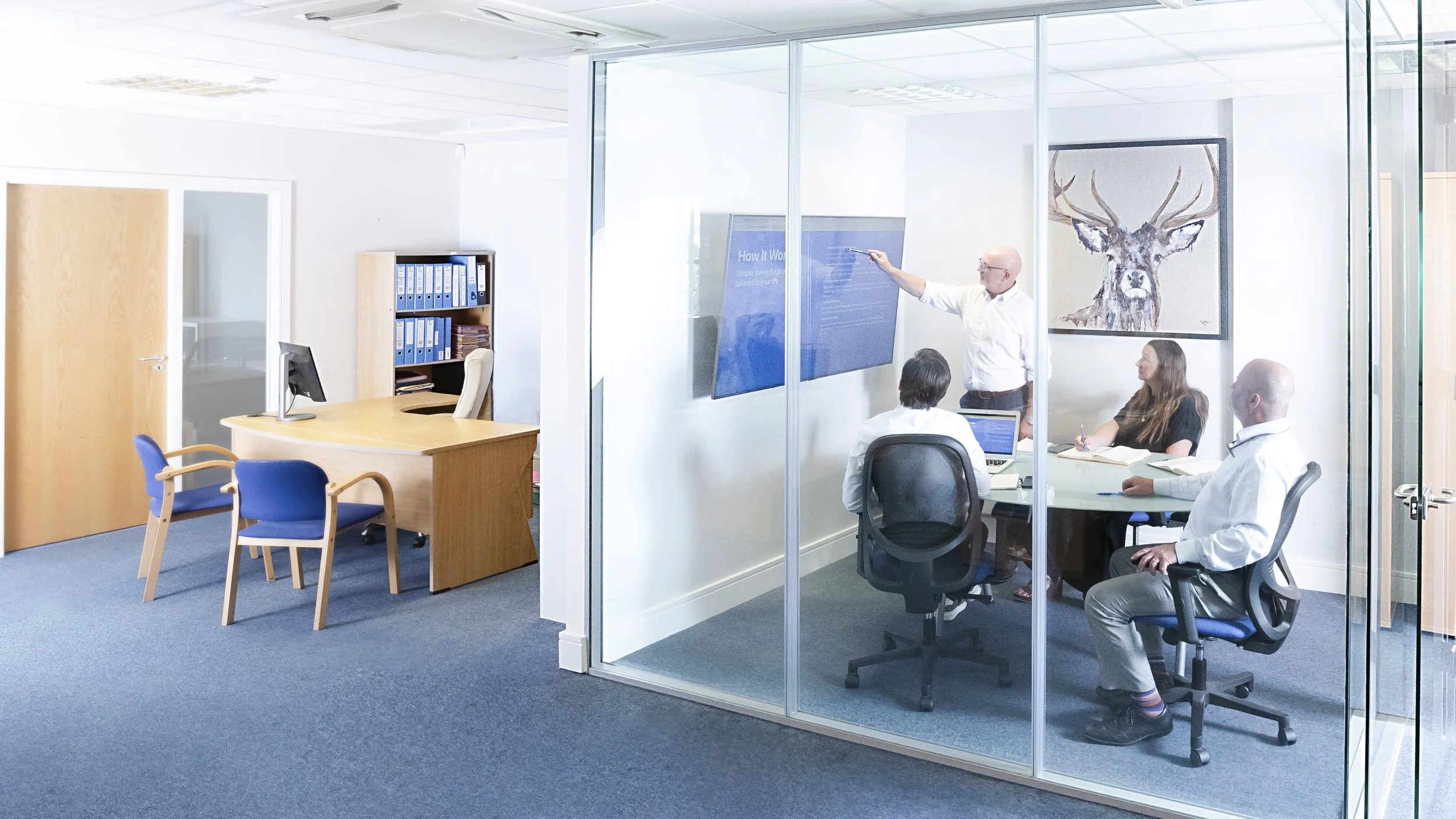 Business meeting with four people in a glass-enclosed conference room. One person is standing and presenting with a touch screen, three are seated, one with a laptop, and two taking notes. There is a large framed deer portrait hanging on the white wall.