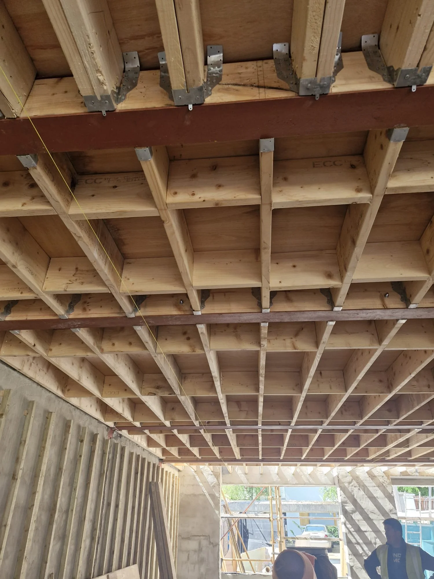 View of a wooden ceiling framework under construction with evenly spaced beams and support brackets, with some workers visible in the background at a construction site.