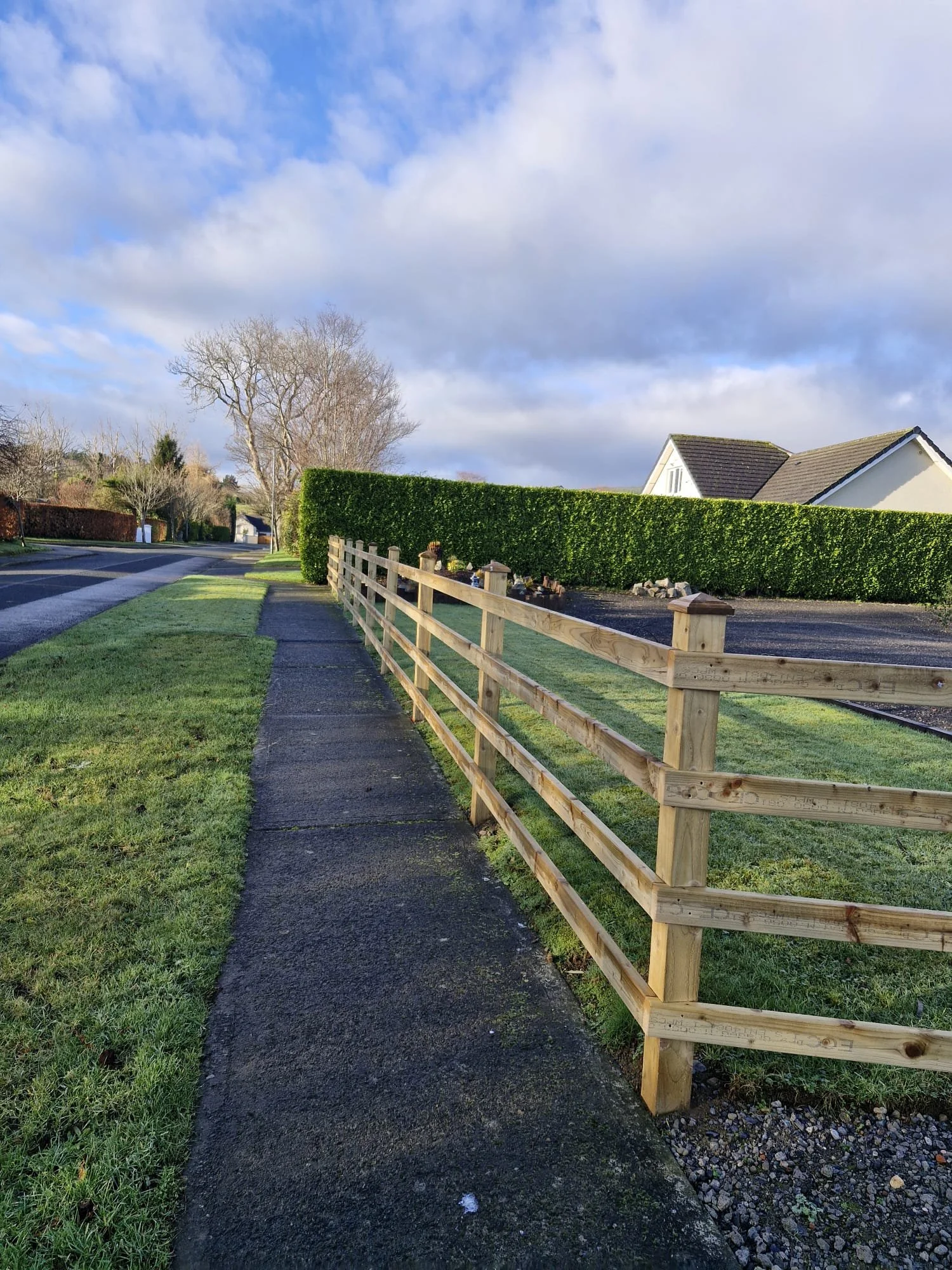 A neighborhood sidewalk with a wooden fence on the right, a hedge, a house with a roof, a tree, and a cloudy sky.
